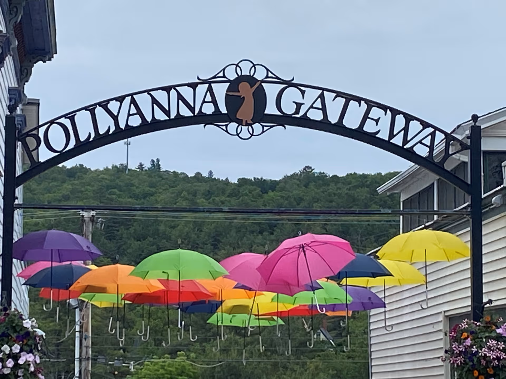 A decorative black metal archway with the words 'Pollyanna Gateway' and a silhouette of a girl in the center. Below the arch, colorful umbrellas in shades of purple, orange, green, pink, yellow, and blue are suspended in the air. In the background, there are green trees and hills under a cloudy sky, with buildings on either side of the street.
