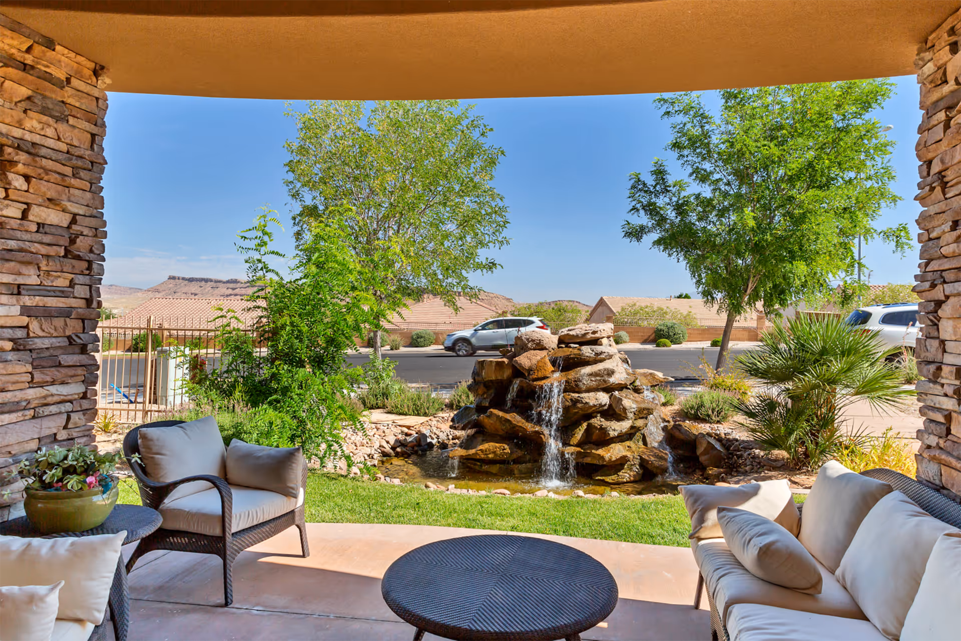 Covered patio with cushioned wicker seating and a round table overlooking a landscaped yard with a rock waterfall.