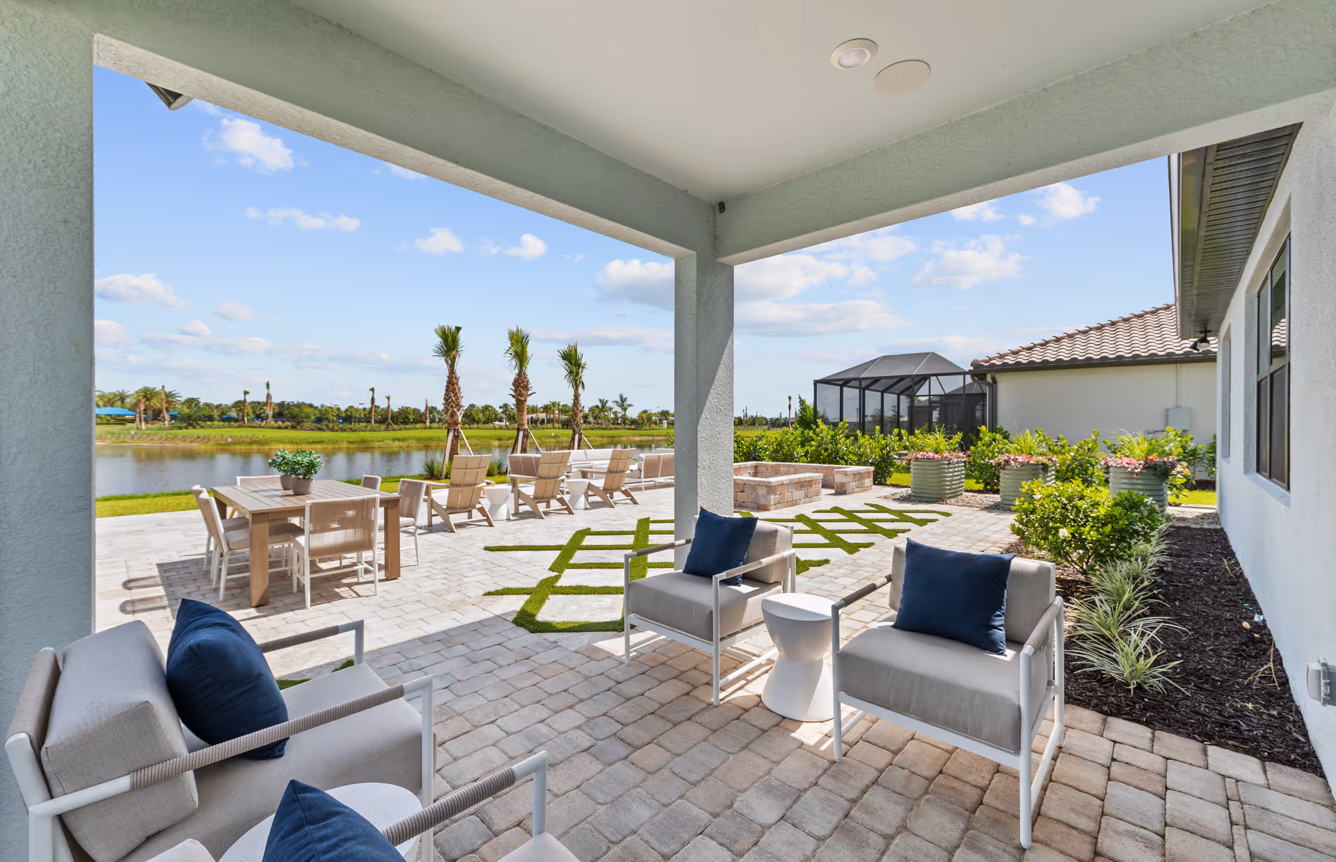 Covered patio area with cushioned chairs and blue pillows overlooking an outdoor space with a dining table, lounge chairs, palm trees, and a water view under a partly cloudy sky.