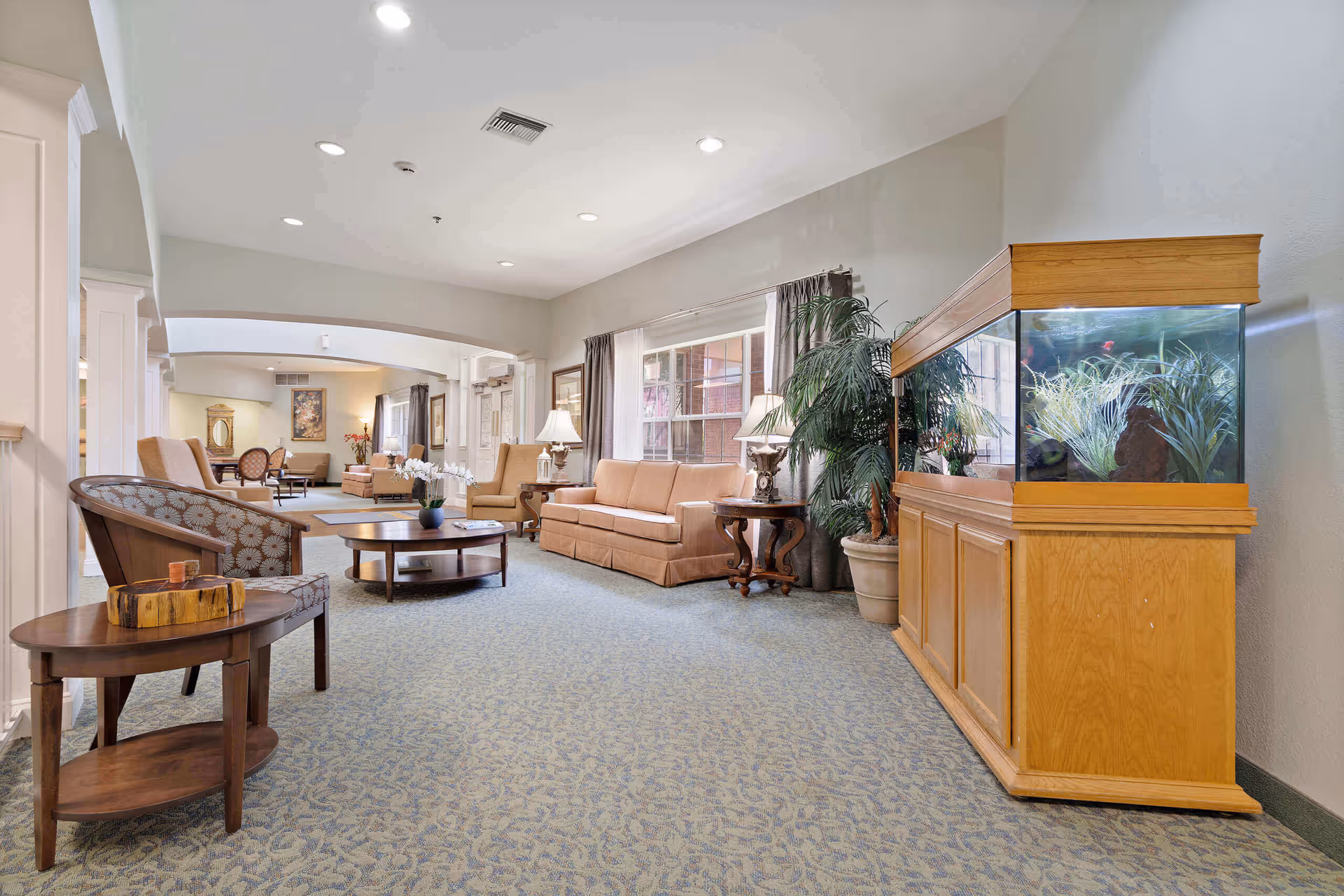 A spacious and well-lit living room area in Morada Cedar Hill featuring beige sofas, armchairs, wooden coffee and side tables, a large fish tank with plants, potted plants, and large windows with curtains allowing natural light to fill the room.