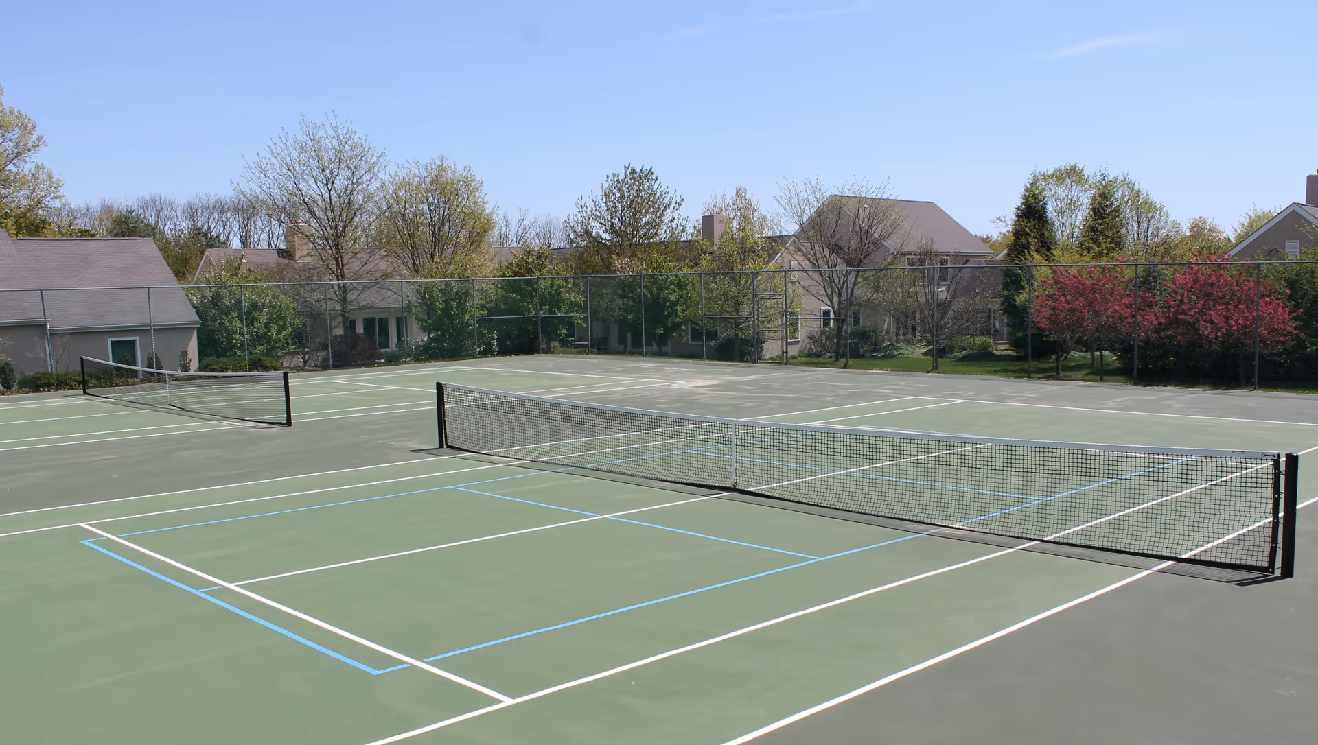 Outdoor tennis court with two nets, surrounded by a chain-link fence and trees with houses visible in the background under a clear blue sky.