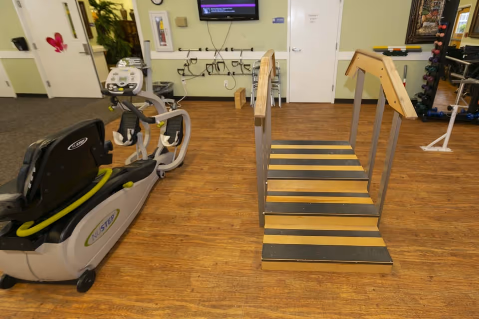 Indoor rehabilitation room with exercise equipment including a NuStep recumbent cross trainer and a set of stairs with handrails for physical therapy. The room has wooden flooring, a wall-mounted TV, and various therapy tools and weights in the background.