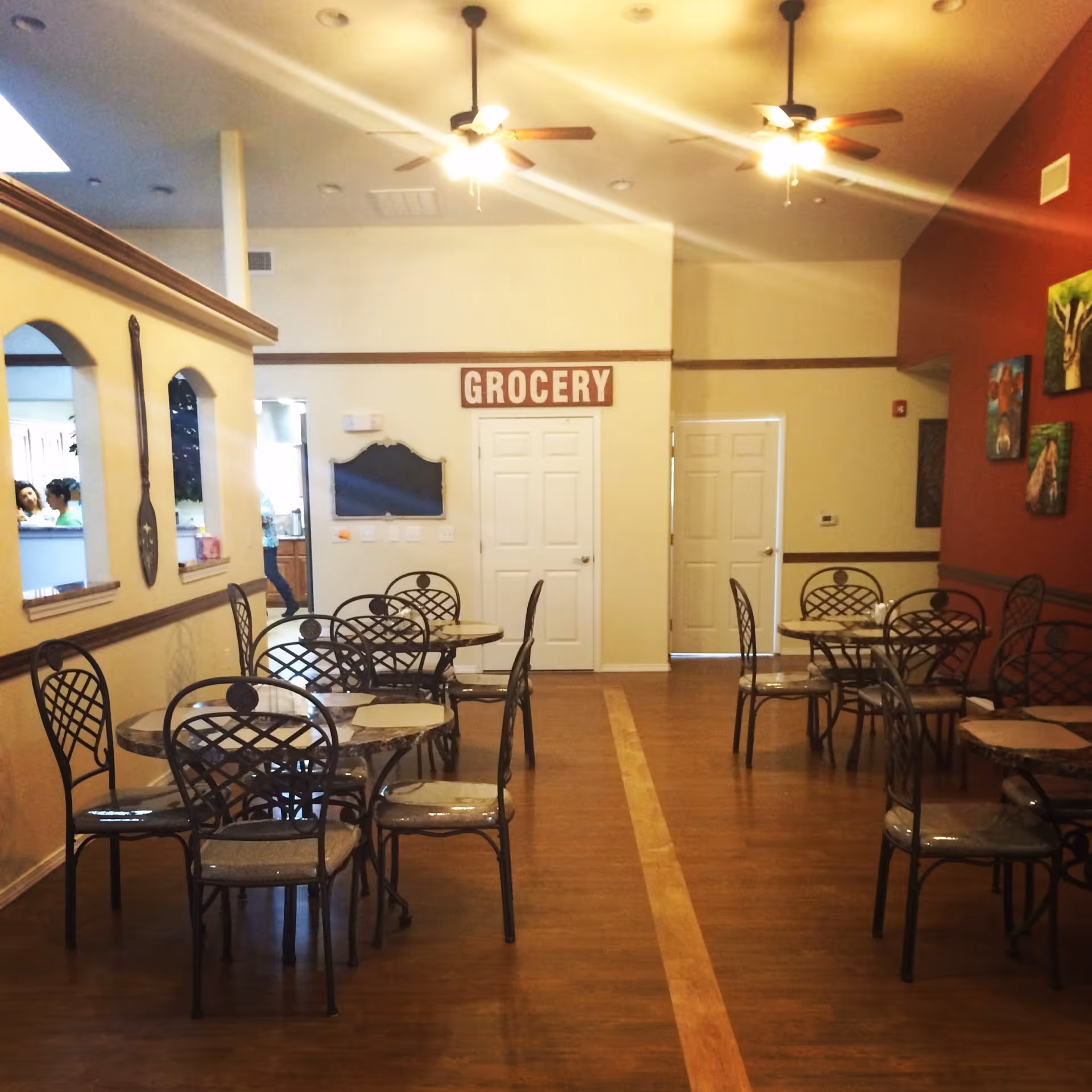 Interior view of a dining area with several round tables and metal chairs with cushions. The room has wooden flooring, ceiling fans with lights, and a red accent wall decorated with paintings. A sign above two white doors reads 'GROCERY'.