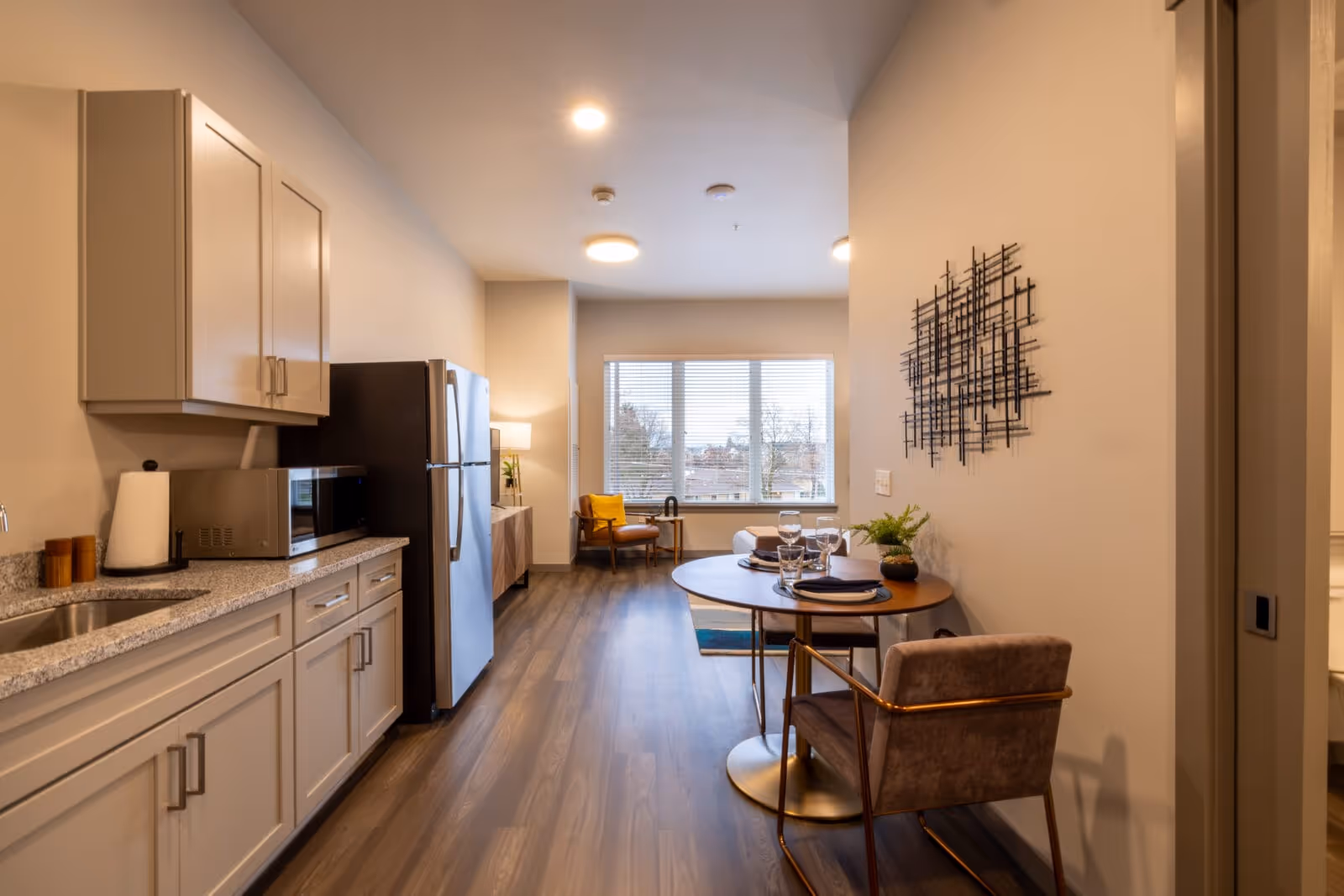 Open-plan senior living apartment interior with a kitchenette on the left, a small dining table and chairs, and a sitting area by a window.