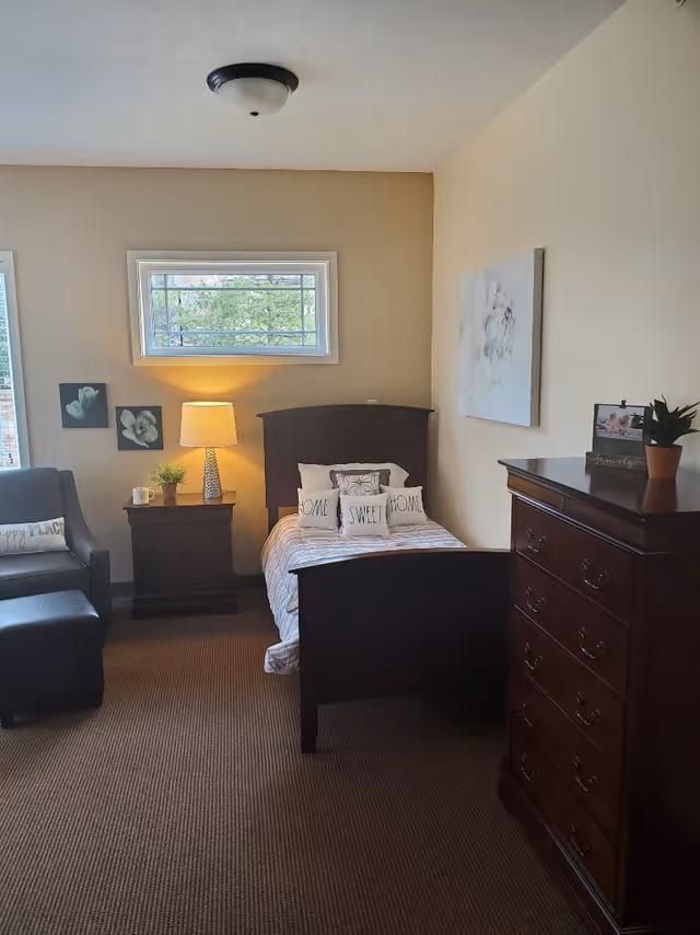 A cozy bedroom with a single dark wood bed featuring pillows that say 'HOME SWEET HOME'. Next to the bed is a nightstand with a lamp, a small plant, and a mug. There is a dark armchair with a pillow that says 'HAPPY PLACE' and a matching ottoman. On the right side, there is a tall dark wood dresser with a small plant and a framed photo on top. The walls are light-colored with floral artwork, and a small window above the bed lets in natural light.