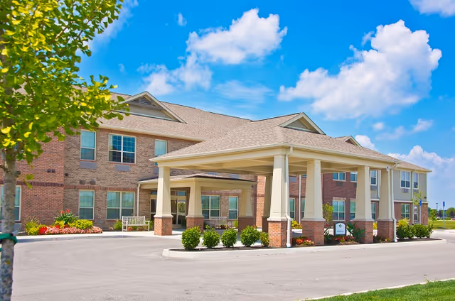 Exterior view of a senior living facility named Entrance 1A - Aspen Trace, showing a large covered entrance with columns, brick and siding exterior, windows, landscaping with bushes and flowers, and a clear blue sky with some clouds.