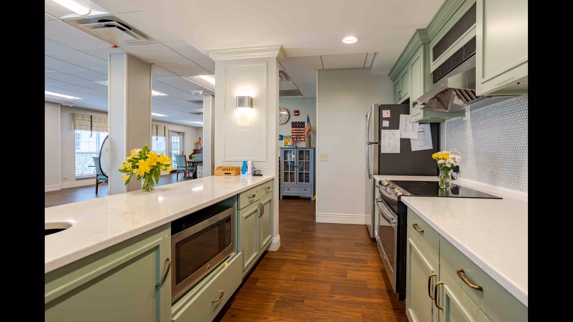 A modern kitchen area with light green cabinets, white countertops, a built-in microwave, stove, and refrigerator. There are vases with yellow flowers on the countertops. The kitchen opens into a common area with large windows and seating visible in the background.