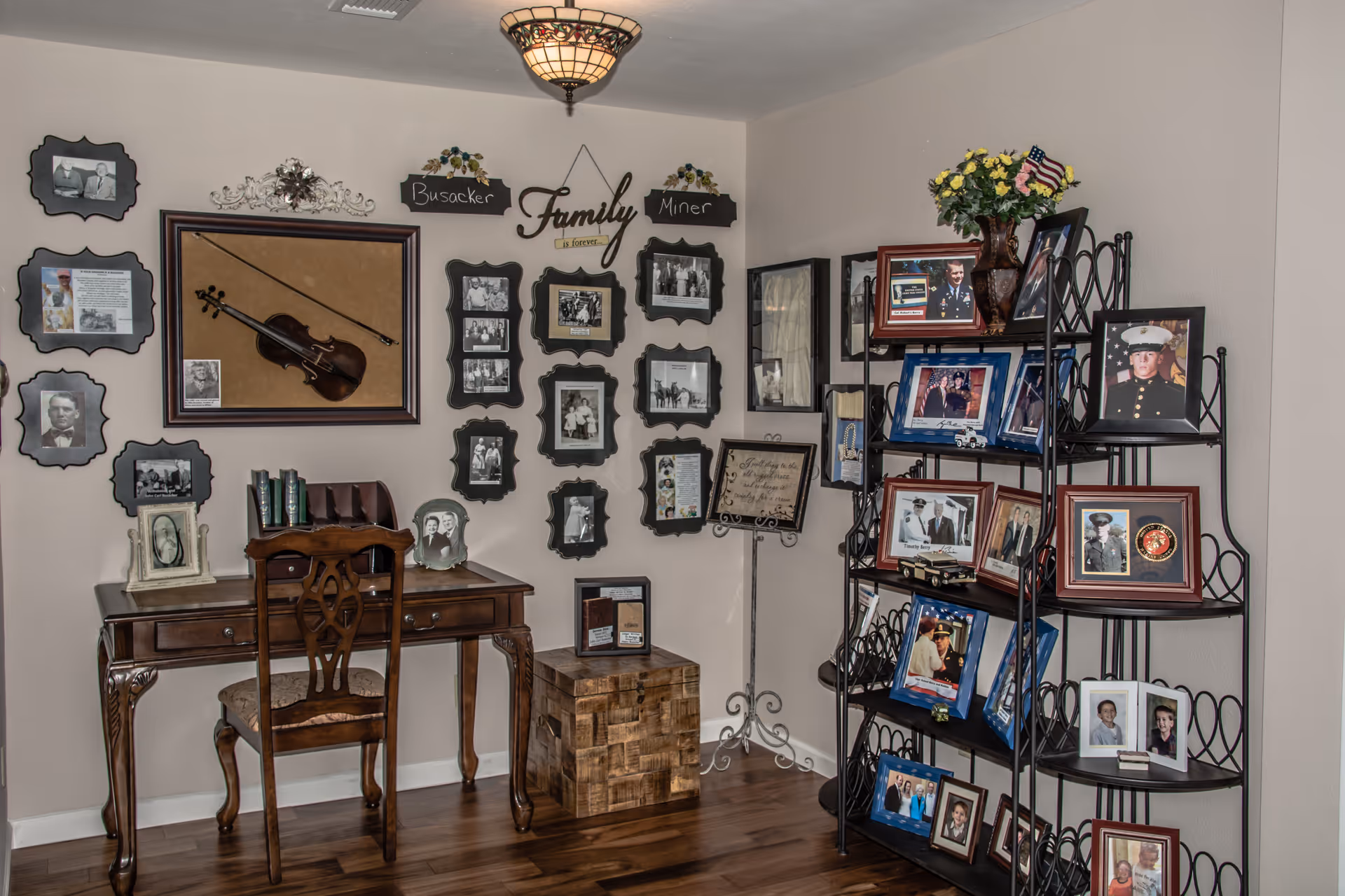 A cozy corner of a room featuring a wooden desk with a chair, a collection of framed family photographs on the walls, and a black metal shelving unit filled with more framed pictures and a vase of flowers. The wall displays a violin in a frame and decorative signs with the words 'Family is forever' and family names.