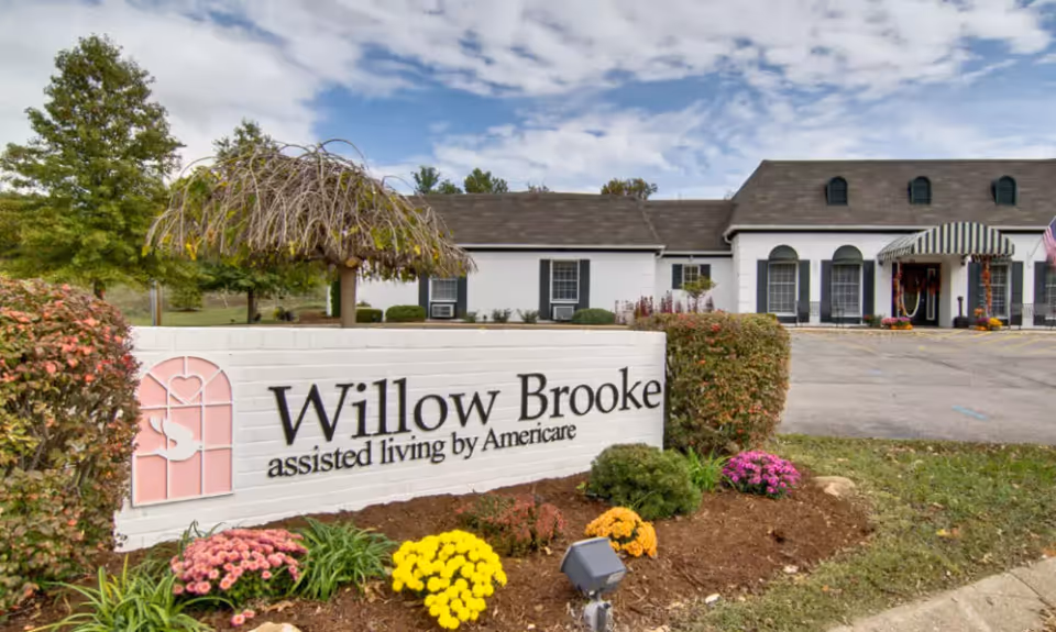 A landscaped entrance sign reading "Willow Brooke assisted living by Americare" in front of a white building with flowers and shrubs.