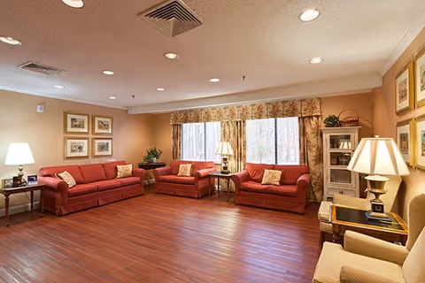 A spacious living room with three red sofas and two beige armchairs arranged around wooden side tables with lamps. The room has wood flooring, beige walls adorned with framed pictures, and large windows covered with floral curtains allowing natural light to enter.