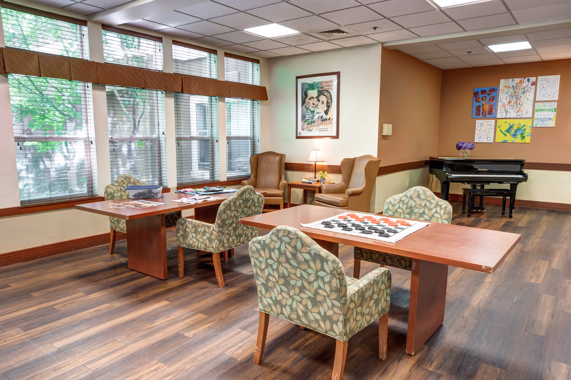 A bright common room with large windows featuring blinds, wooden floors, and two tables with patterned chairs. One table has a checkers game set up, and the other has puzzles and games. In the corner, there are two brown armchairs with a small table and lamp between them. A piano is against the far wall, which is decorated with colorful artwork and a framed Casablanca movie poster.
