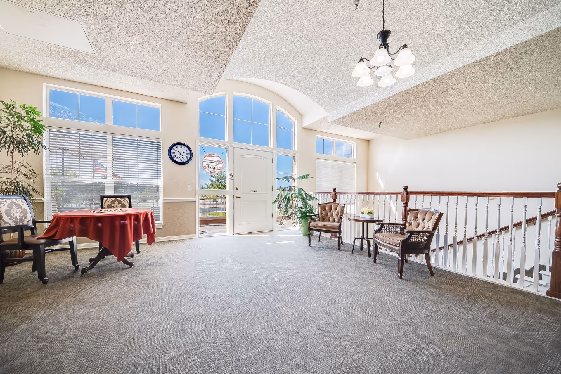 Bright assisted-living lobby with large arched windows, seating areas, a small table with a red tablecloth, potted plants, and a staircase railing.