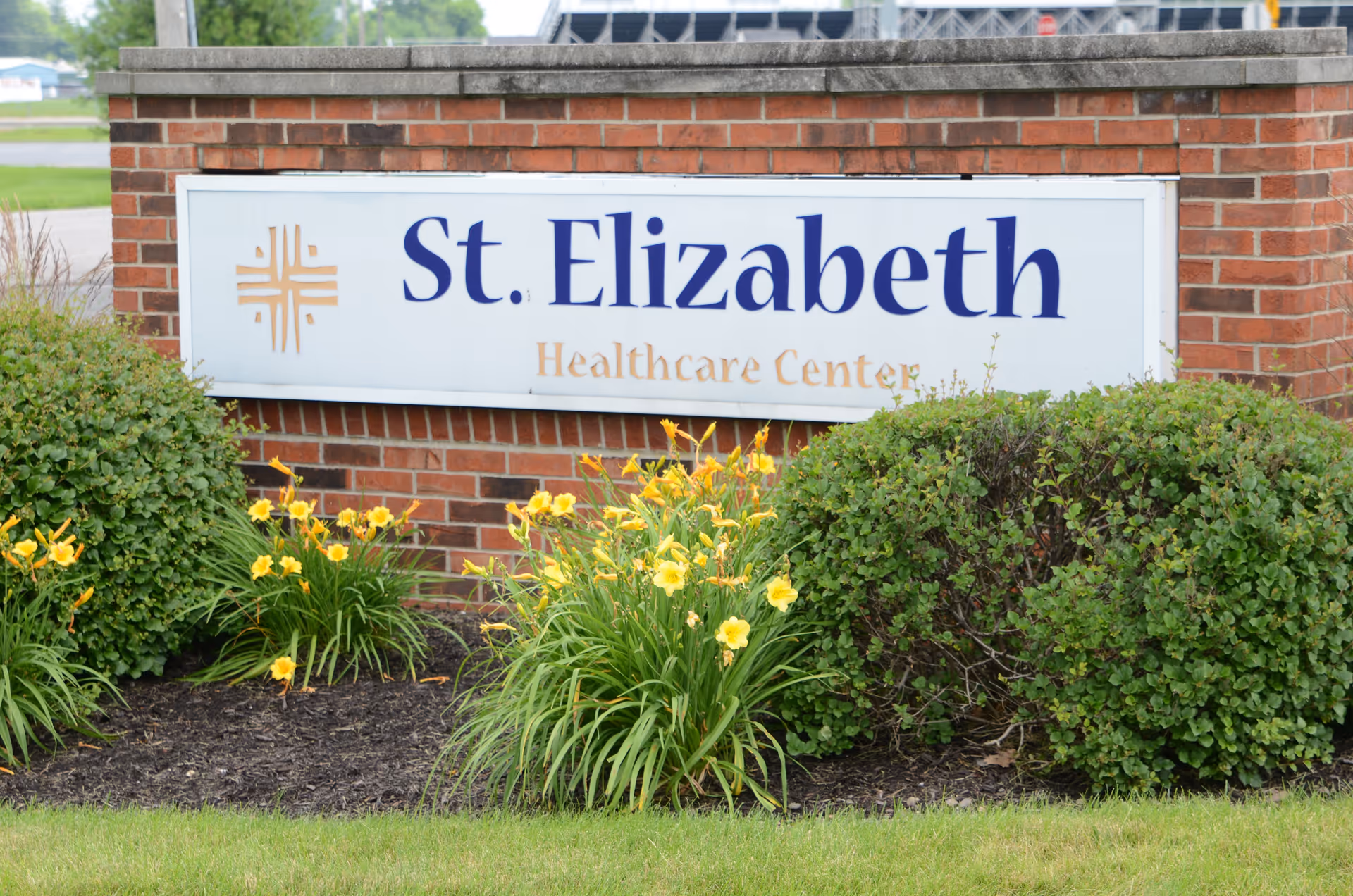 Brick entrance sign reading 'St. Elizabeth Healthcare Center' with shrubs and yellow flowers in front.