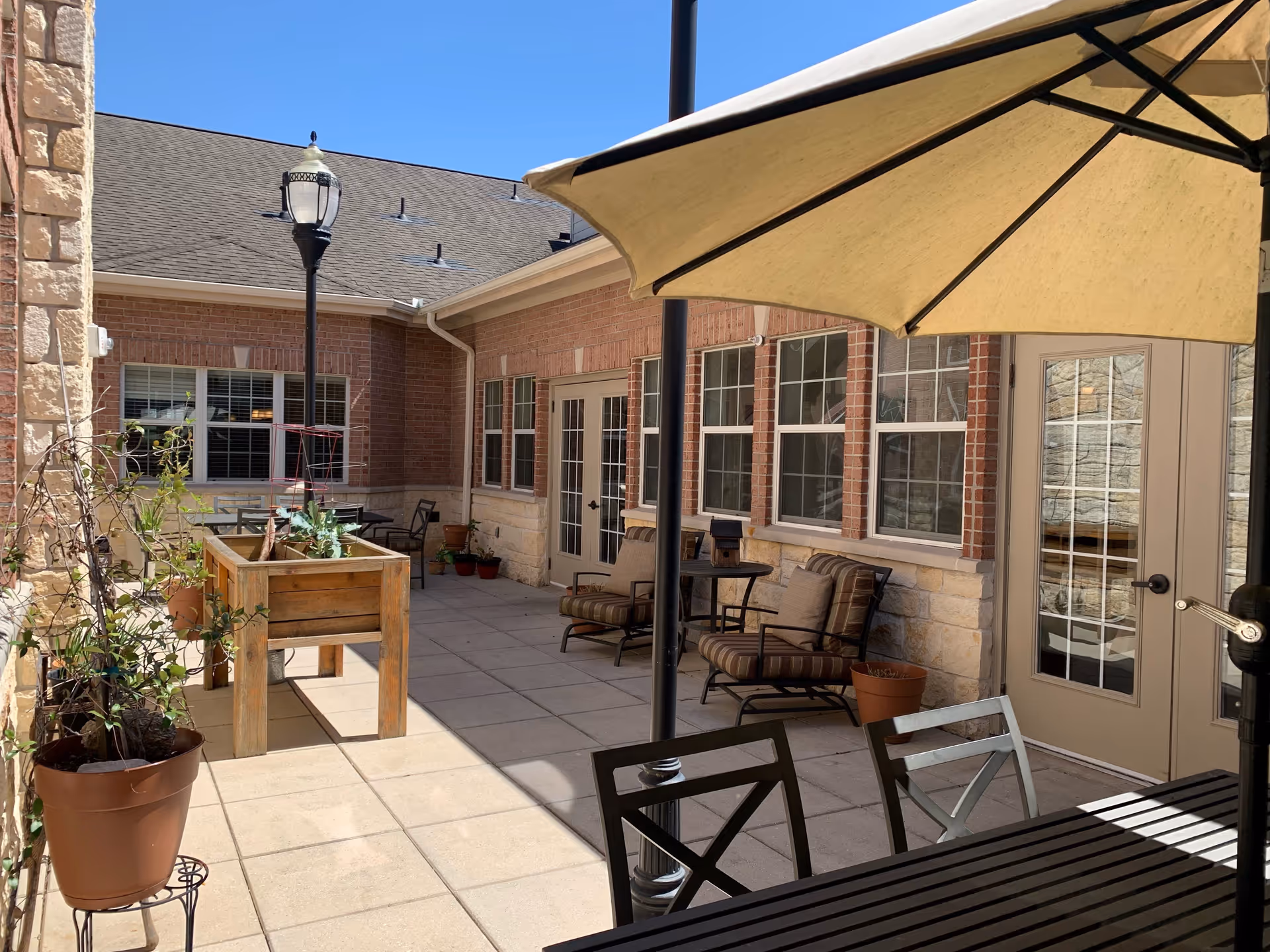 Outdoor patio area at Creekside Alzheimer's Special Care Center with a large beige umbrella shading a table and chairs. There are potted plants, a raised wooden planter box, cushioned chairs, and a lamppost. The building has brick and stone walls with multiple windows and glass doors.