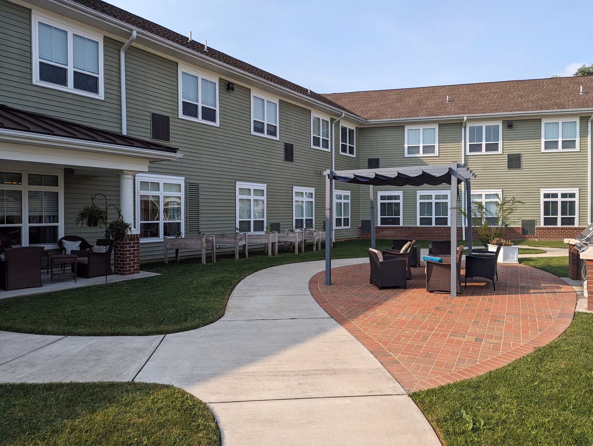 Outdoor courtyard area of a senior living facility with a paved walkway, green grass, and a seating area under a pergola. The building has green siding with white trim and multiple windows. There are chairs and a table under the pergola and additional seating on a covered porch to the left.