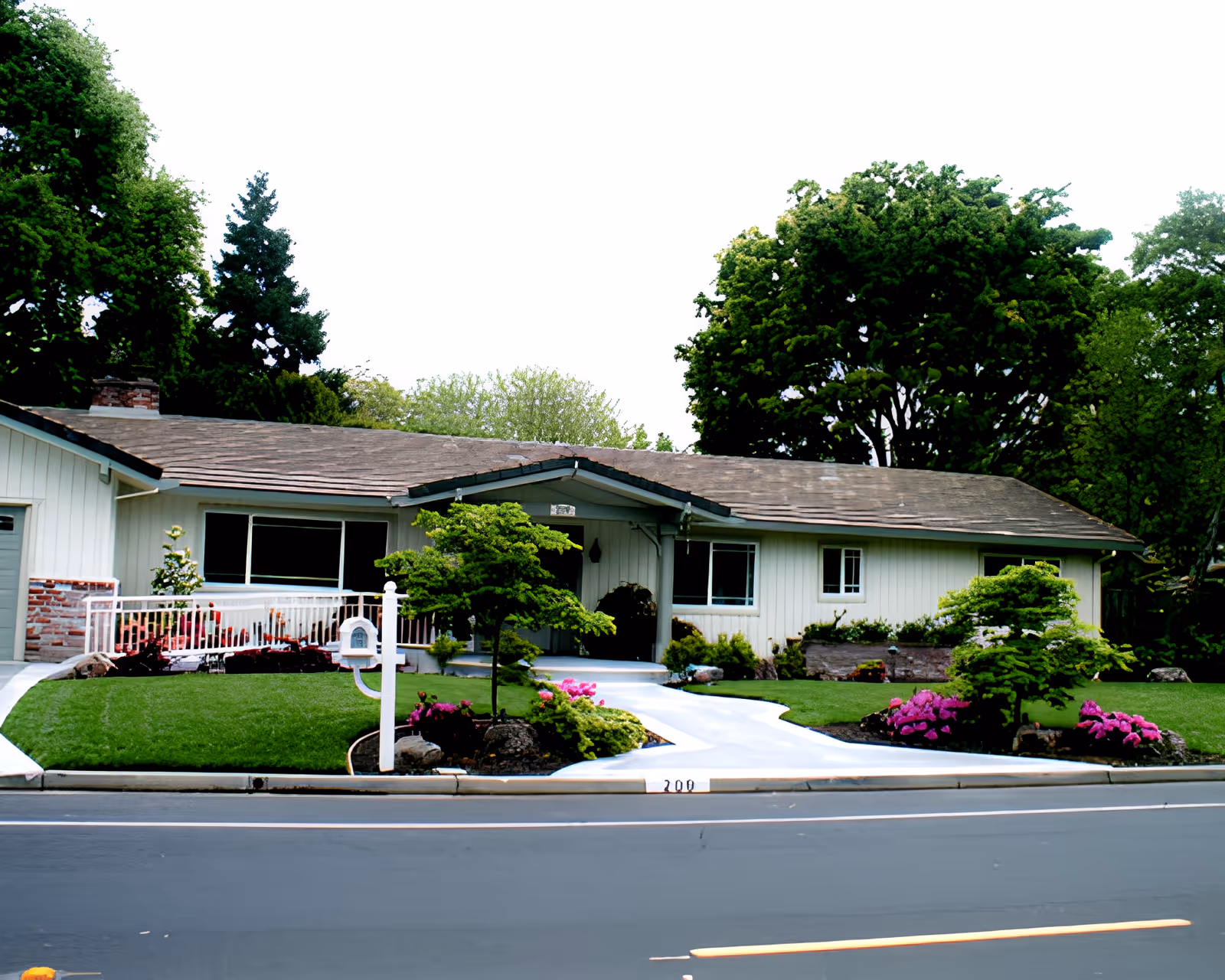 Single-story residential building with a sloped roof, surrounded by green trees and landscaped garden with bushes and flowers. A white mailbox stands near the sidewalk, and a paved walkway leads to the front door.