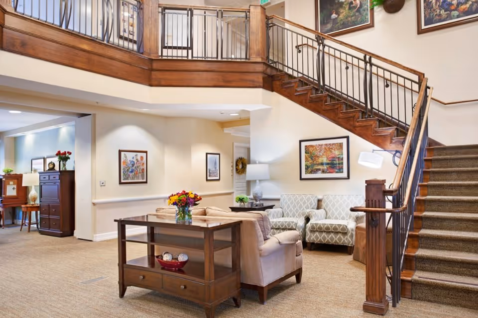 Interior view of a senior living facility lounge area with a staircase. The room features comfortable seating including armchairs and a sofa, a wooden side table with a vase of flowers, framed artwork on the walls, and warm lighting. The staircase has wooden handrails and metal balusters, leading to an upper floor with a railing.