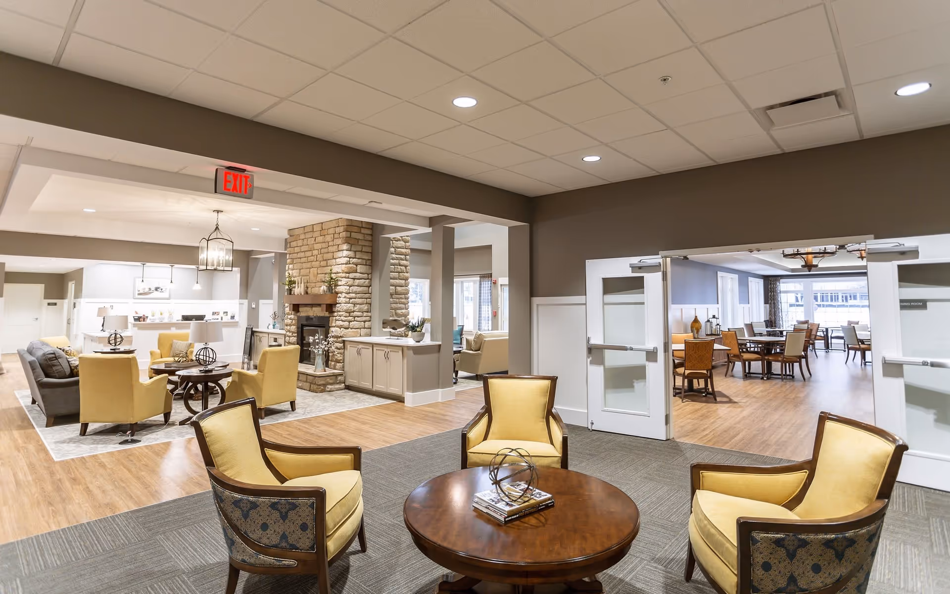 A spacious and well-lit common area in a senior living facility featuring a seating arrangement with four yellow upholstered chairs around a round wooden table with decorative items on it. In the background, there is a cozy sitting area with a stone fireplace, additional seating, and a dining room visible through double doors. The flooring is a combination of carpet and wood, and the ceiling has recessed lighting.