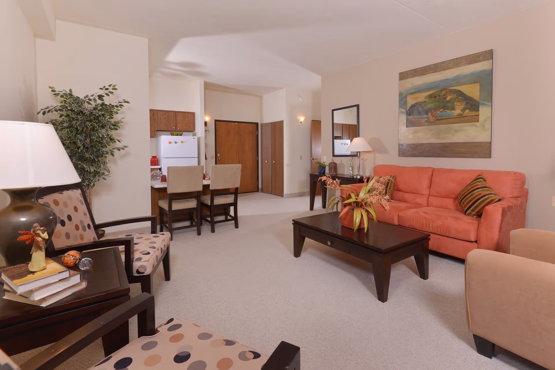 A cozy senior living room featuring a coral-colored sofa with striped pillows, a dark wooden coffee table with a plant centerpiece, two patterned armchairs, a side table with books and a lamp, and a small dining area with two chairs near a white refrigerator. The walls are decorated with a large painting and a mirror, and there is a potted plant in the corner.