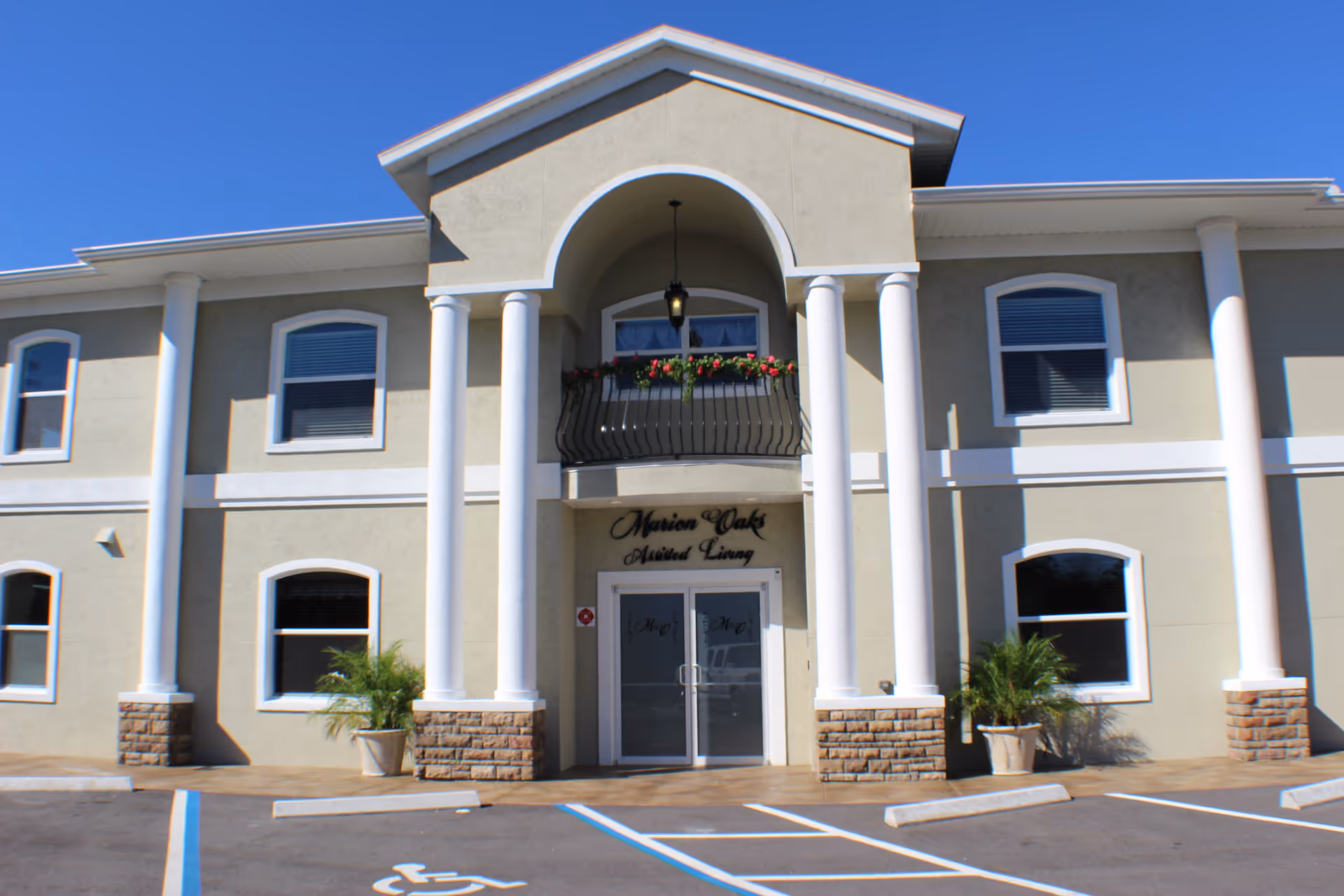 Front exterior view of Marion Oaks Assisted Living building with beige walls, white columns, arched entrance, and a small balcony with flowers above the entrance. There are several windows and a parking area with a handicapped parking space in front.