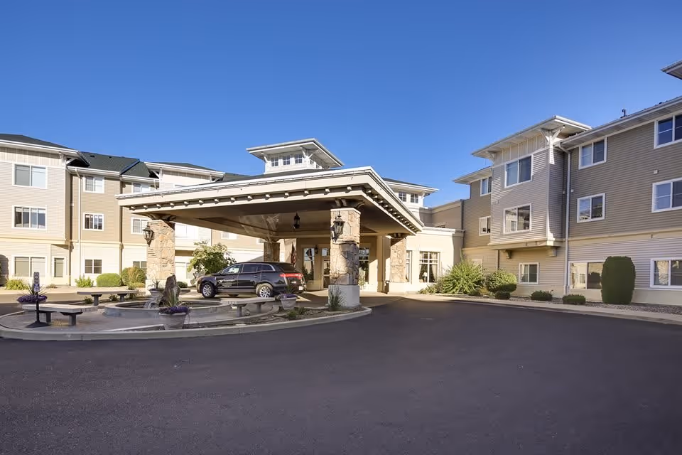 Exterior view of Fairwinds - Spokane senior living facility showing a large covered entrance with stone pillars, a black SUV parked under the canopy, and a circular driveway with benches and planters. The building is three stories tall with beige siding and multiple windows under a clear blue sky.