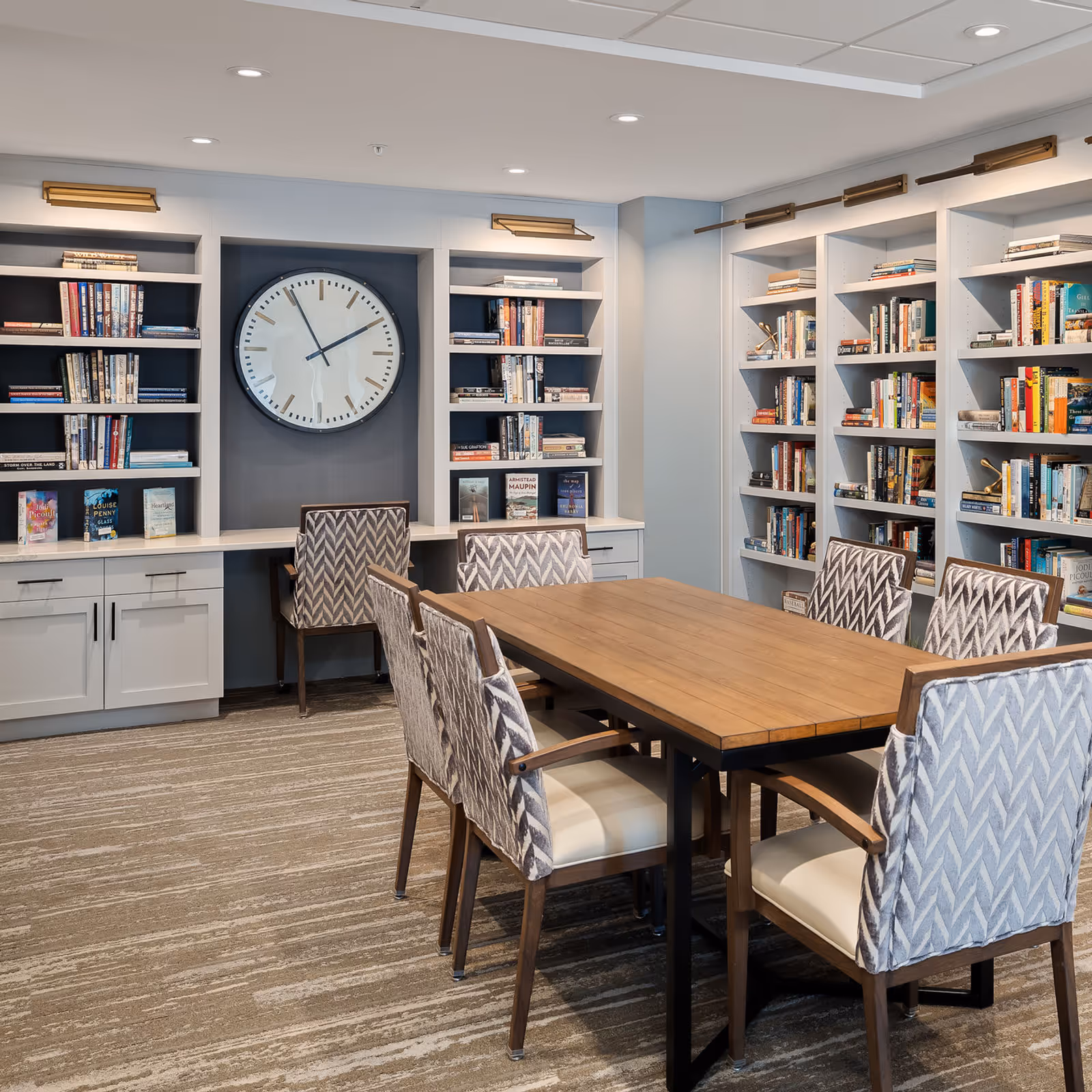 A well-lit communal library/meeting room with a wooden table surrounded by patterned upholstered chairs, built-in bookshelves, and a large wall clock.