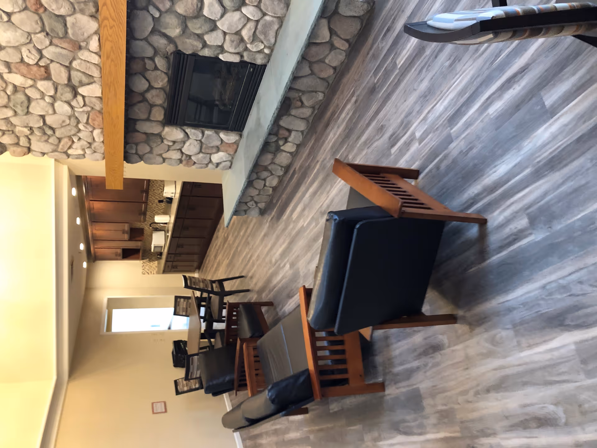 Community lounge with wooden-framed chairs and leather armchairs facing a stone fireplace and a kitchenette in the background.