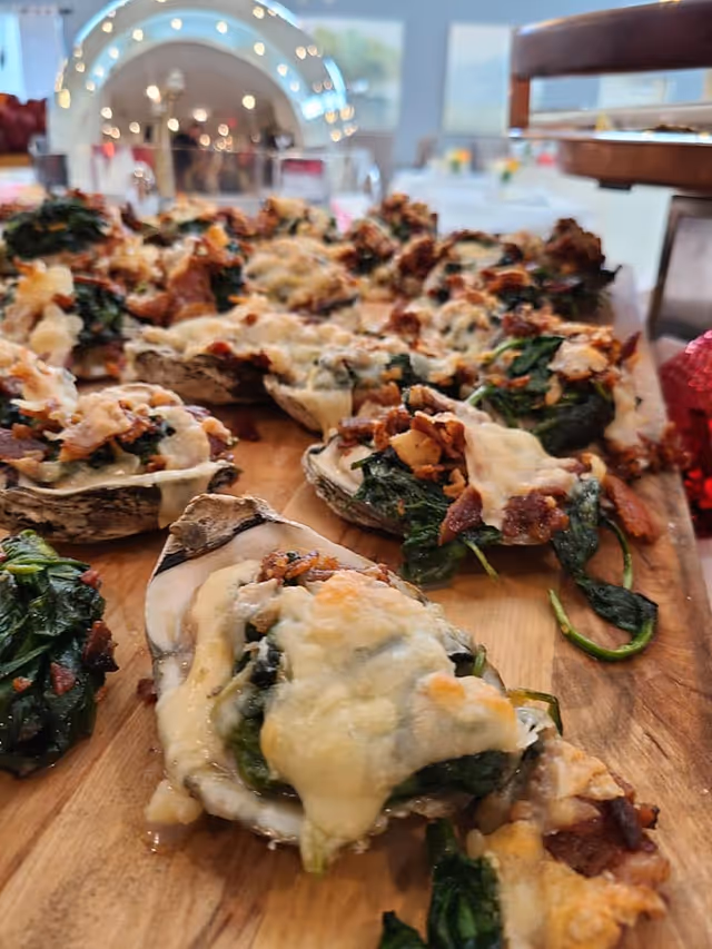Close-up view of baked oysters topped with spinach, melted cheese, and bacon bits on a wooden serving board, with a blurred background showing a dining area with tables and chairs.