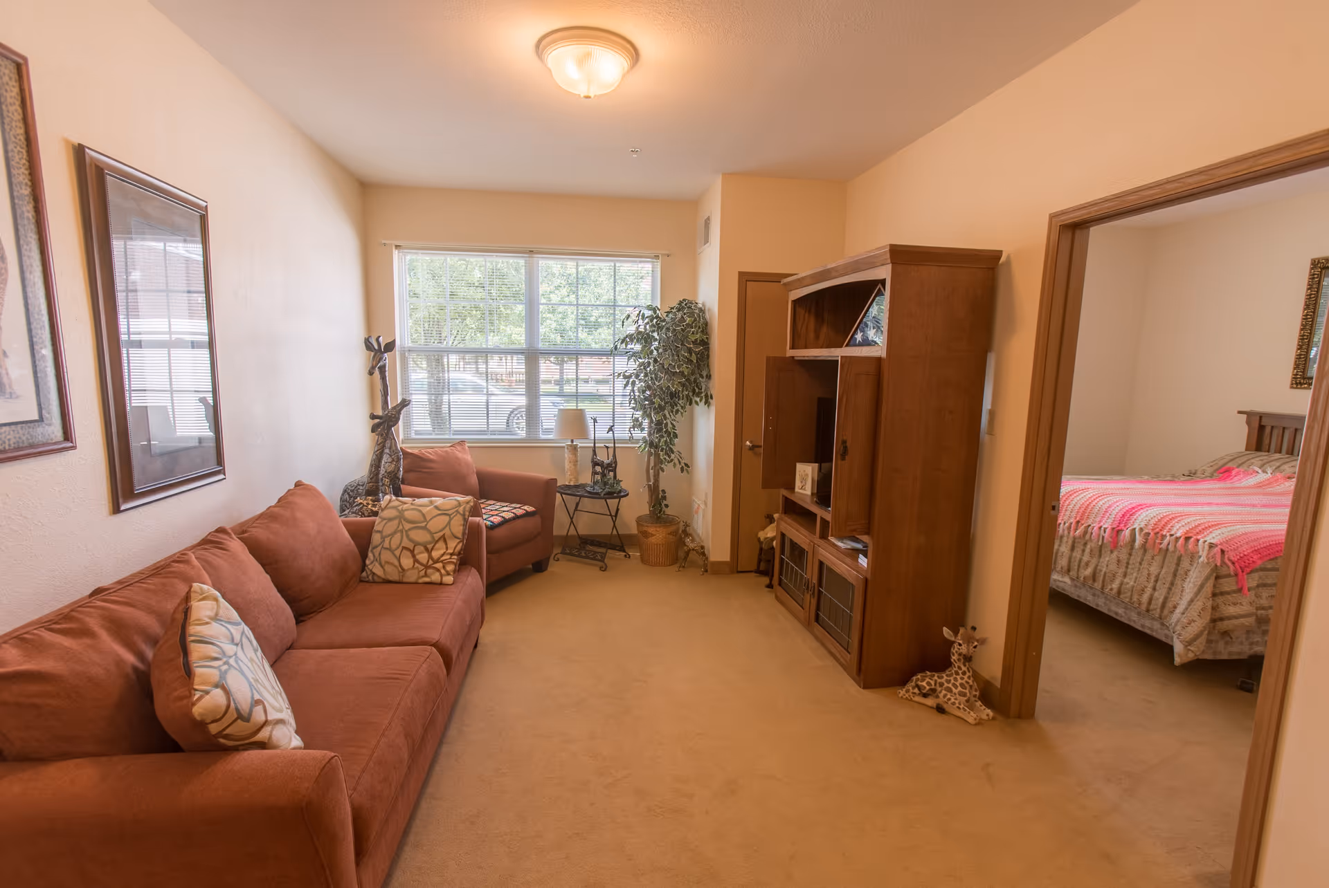 A cozy living room with a rust-colored sofa and matching armchair, decorative giraffe statues, a wooden entertainment center, and a window letting in natural light. Adjacent to the living room is a bedroom with a bed covered in a pink and white blanket.