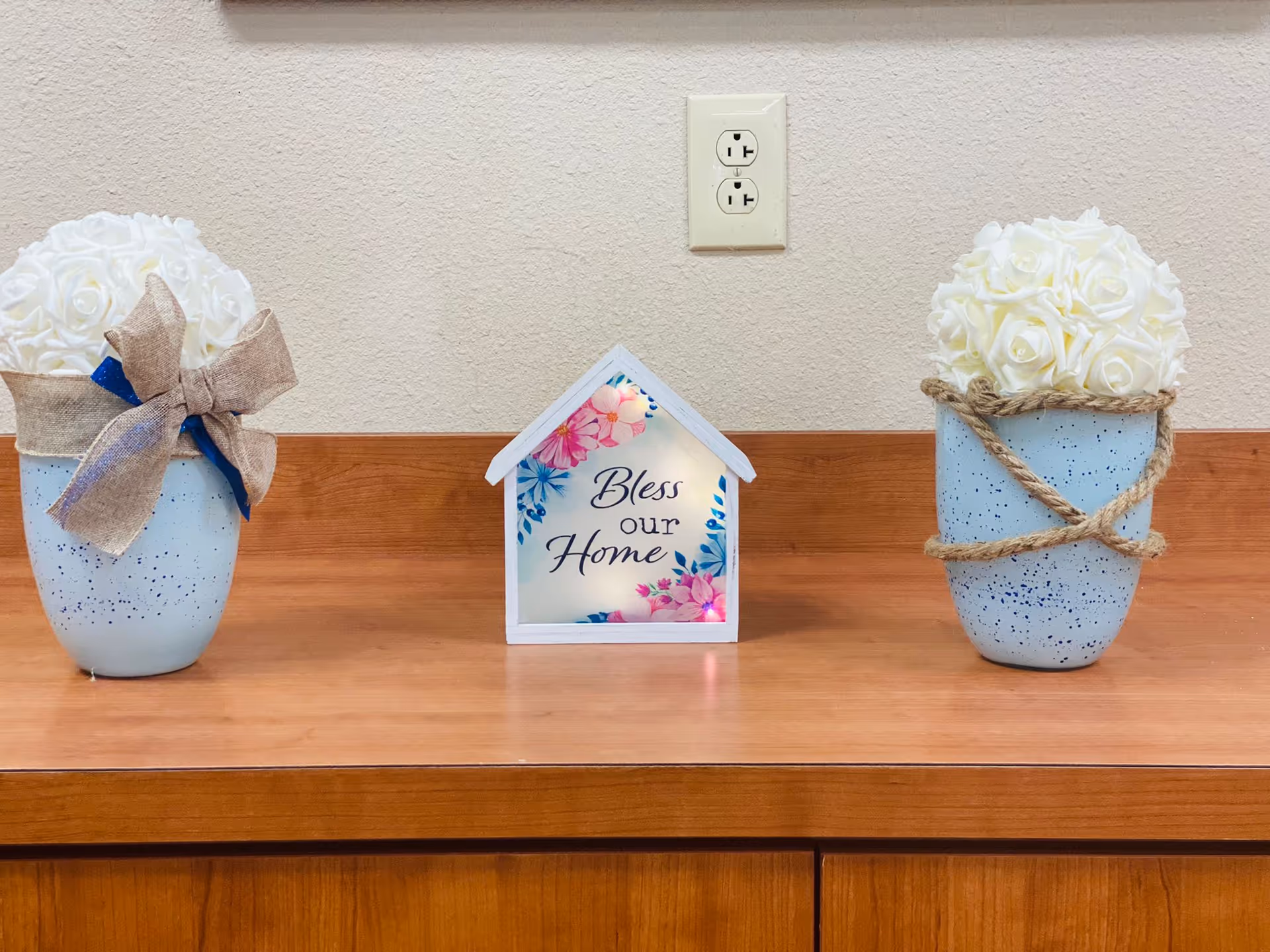 Wood countertop with a small decorative house sign reading 'Bless our Home' flanked by two vases of white artificial roses.