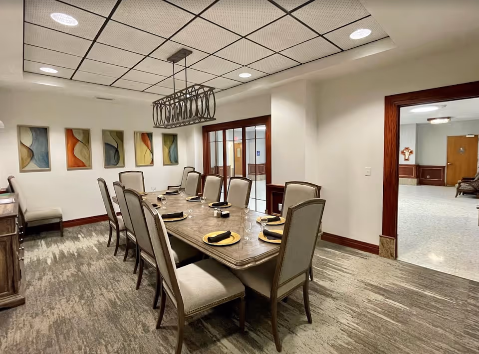 A formal dining room with a long table set for eight, upholstered chairs, a modern chandelier, and abstract wall art.