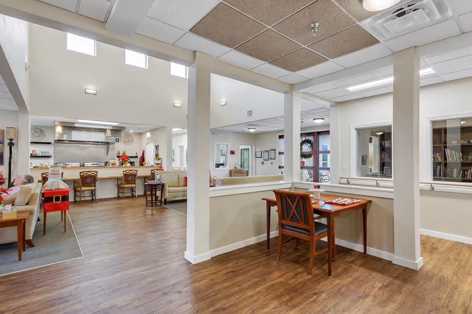 Interior view of a senior living facility common area with wooden flooring, a small table with a chair, a seating area with sofas, and a kitchen area with bar stools and stainless steel appliances. The space is well-lit with natural light from high windows and ceiling lights.