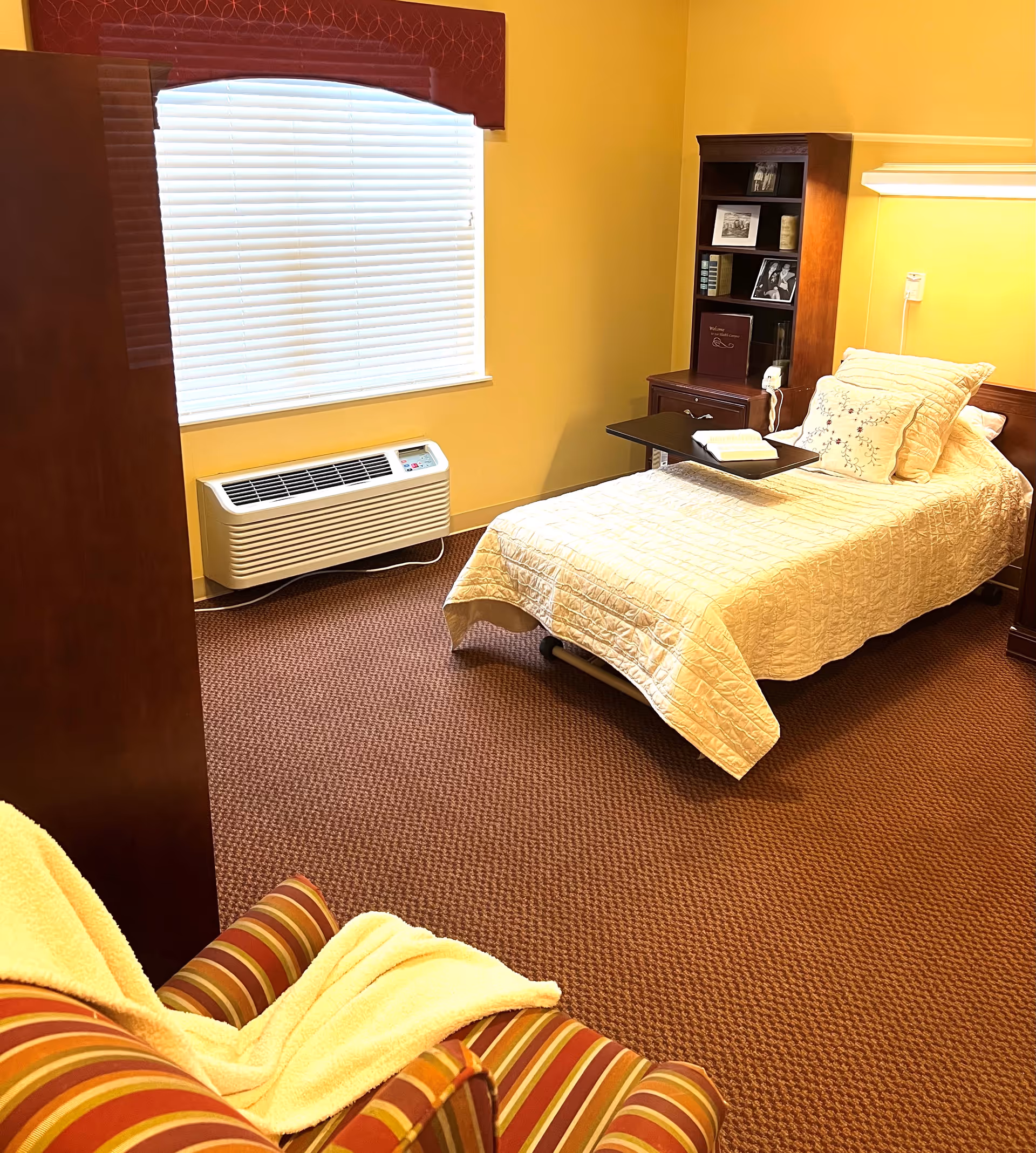 Single-occupancy furnished bedroom with a bed, overbed table, bookshelf nightstand, striped armchair and a window air conditioner.