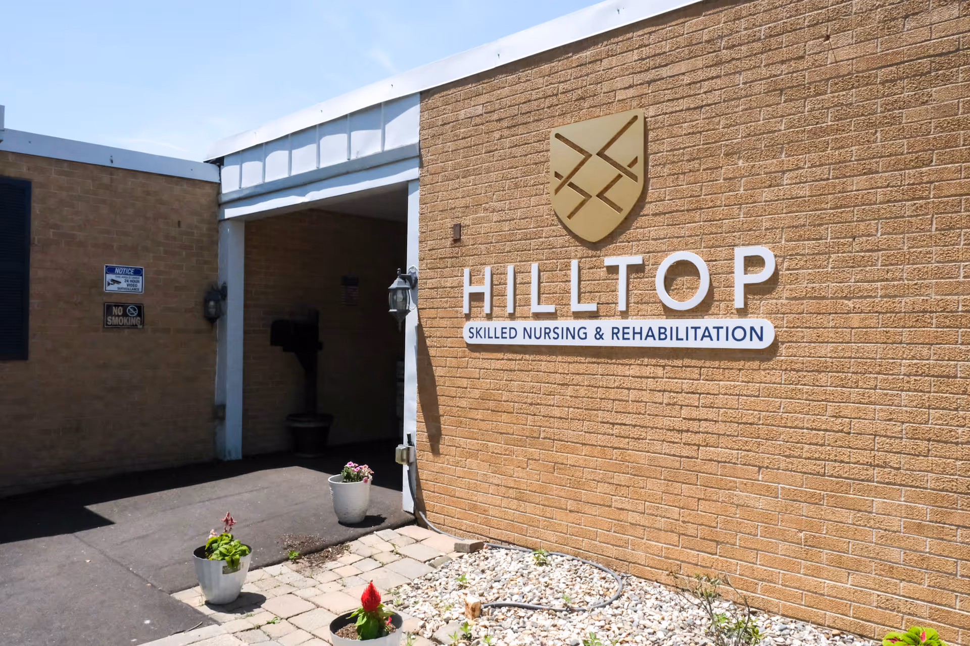 Entrance of Hilltop Skilled Nursing & Rehabilitation showing a brick exterior wall with a large sign, covered entryway, and potted plants.