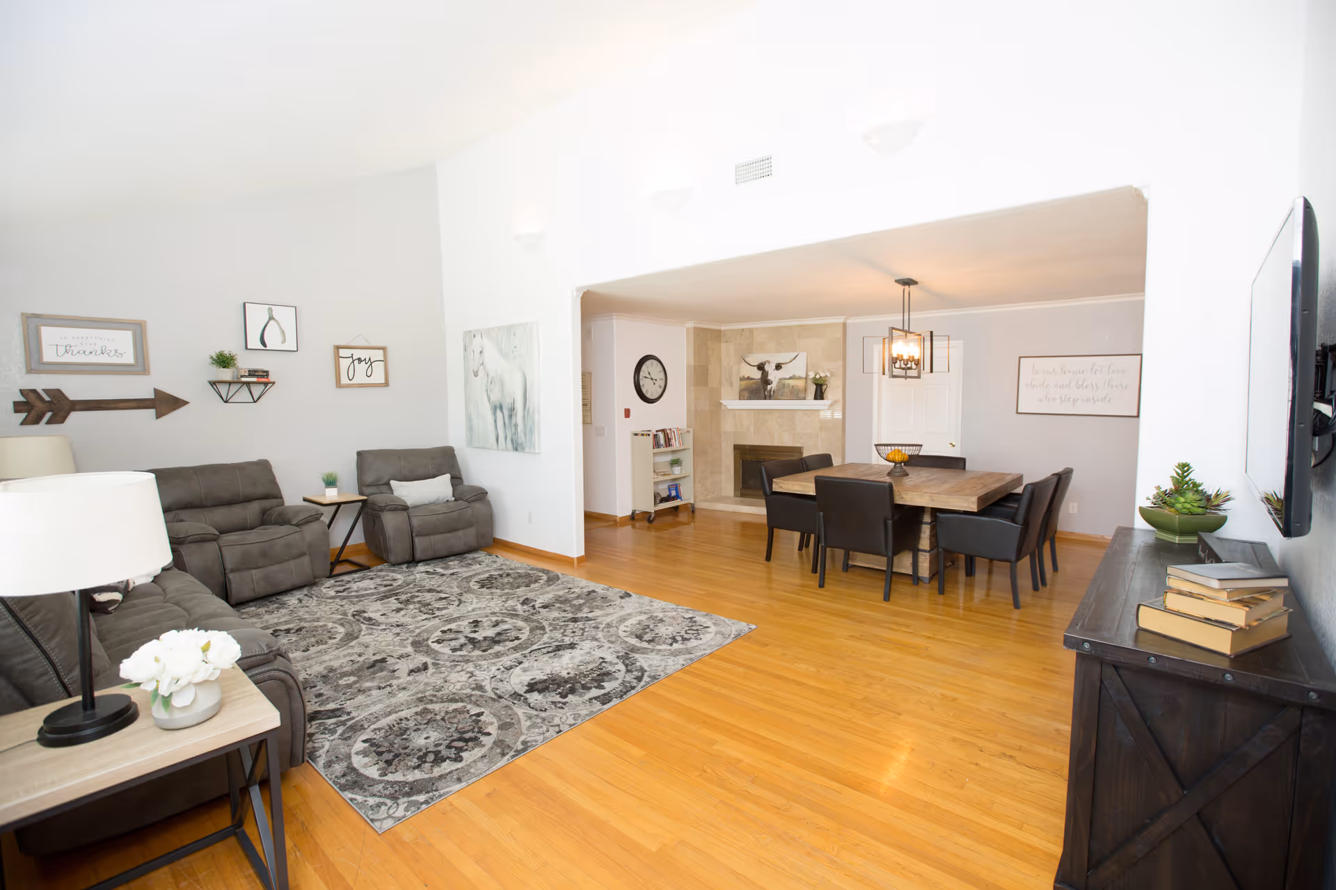 A bright and spacious living and dining area with wooden flooring. The living room features a gray sofa, two matching recliners, a patterned area rug, and wall decorations including framed quotes and artwork. The dining area has a wooden table with six black chairs, a modern chandelier, and a fireplace with a painting above it. A dark wooden console with books and a plant is positioned under a wall-mounted TV.