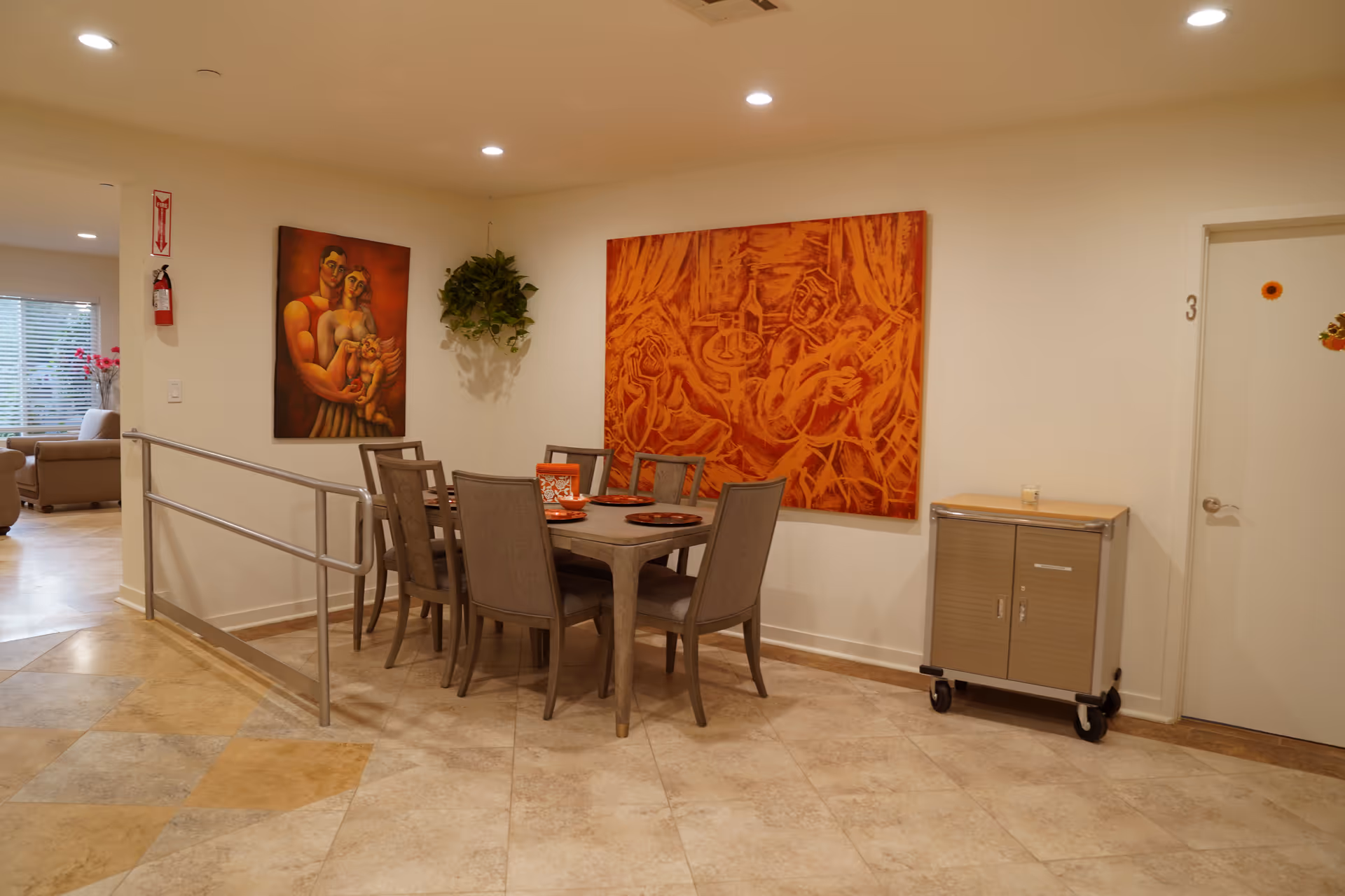 A dining area with a rectangular table surrounded by six chairs. The table is set with orange plates and a decorative box. Two large orange-themed paintings hang on the walls, one depicting a couple with a child and the other showing musicians. A small cabinet on wheels is positioned against the wall near a door labeled with the number 3. The floor is tiled with a beige and light brown pattern, and a metal handrail is installed along the left side of the dining area. In the background, a living room with sofas and a window with blinds is partially visible.