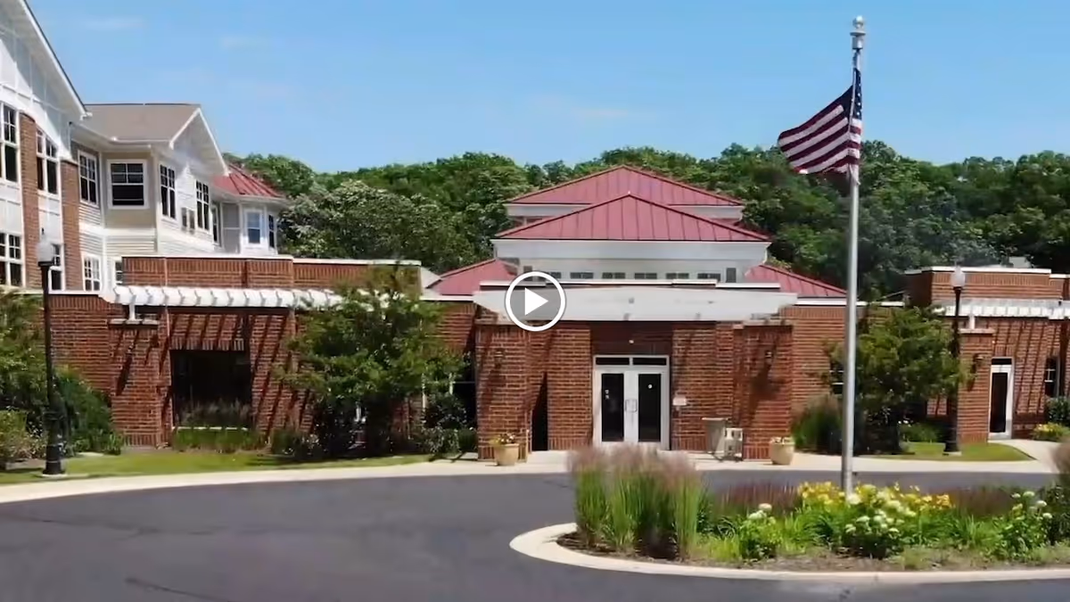 Front exterior of a brick senior living facility with a red-roofed entrance, circular driveway, landscaping, and an American flagpole.
