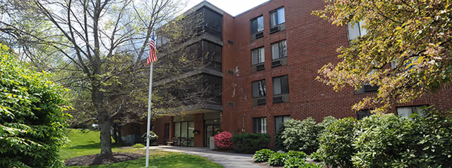 Exterior view of a multi-story brick building surrounded by green shrubs and trees, with a flagpole displaying the American flag in front of the entrance.