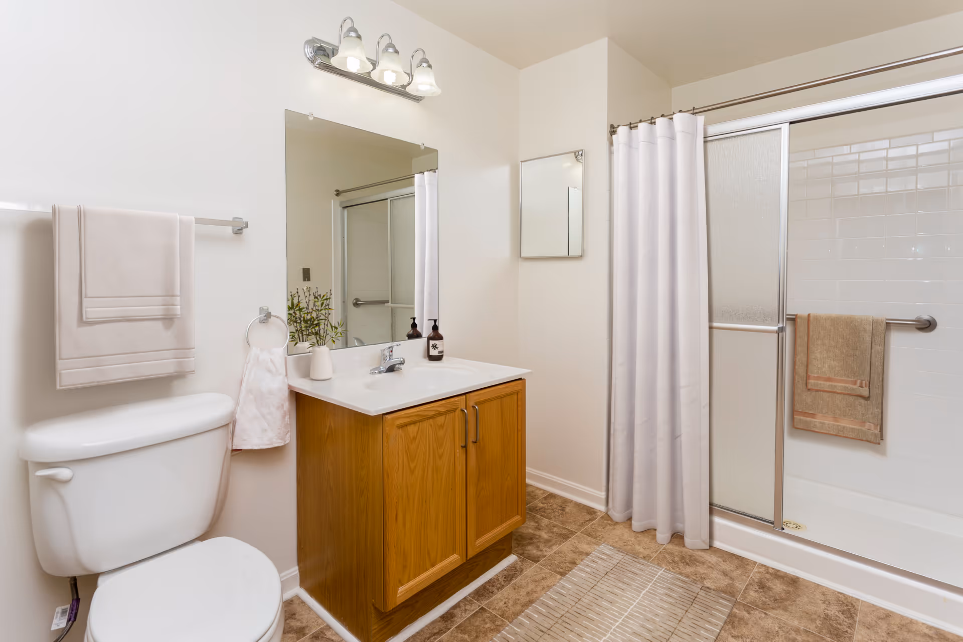 Bright bathroom with a toilet, wooden vanity and sink under a mirror, and a shower with sliding glass doors and curtain.