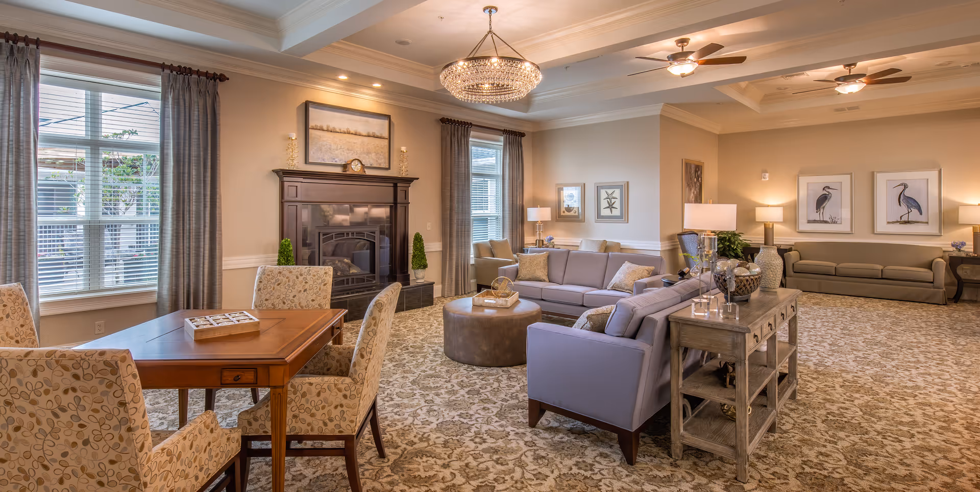 A spacious and elegantly decorated living room with beige walls and patterned carpet. The room features a wooden table with four upholstered chairs, a large fireplace with a dark wooden mantel, two large windows with gray curtains, two gray sofas arranged around a round ottoman, several table lamps, framed artwork on the walls, and ceiling fans with lights.