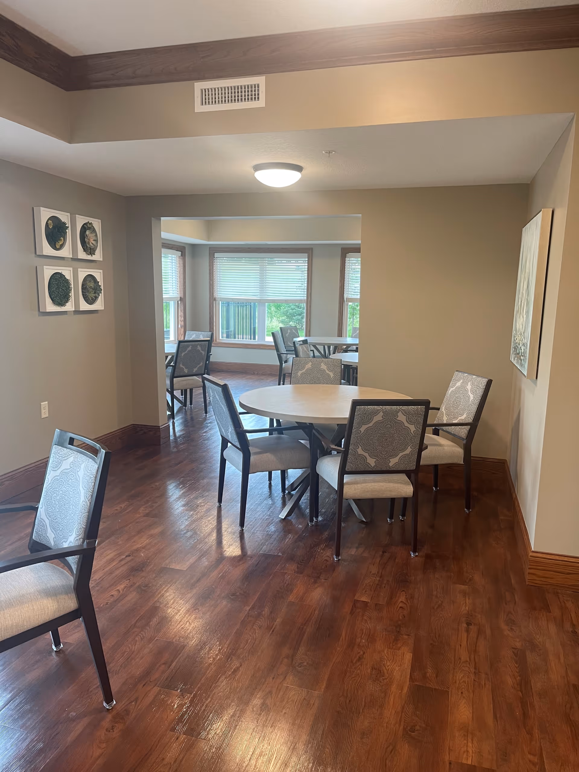 Interior view of a senior living facility dining area with round tables and cushioned chairs on a wooden floor. The room has beige walls, large windows with blinds, and decorative wall art. Natural light brightens the space.