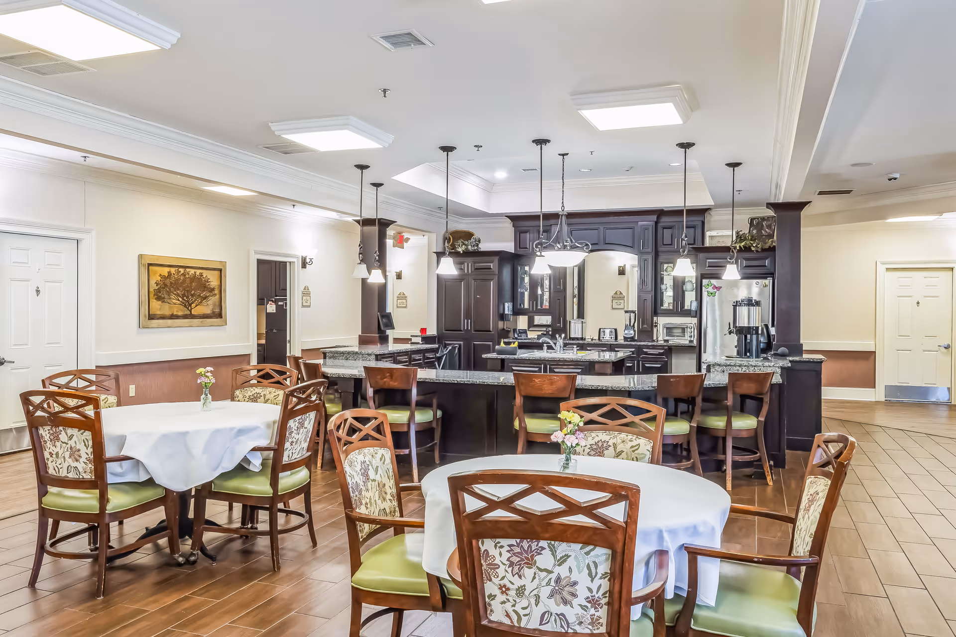 A spacious dining area in a senior living facility featuring round tables with white tablecloths and floral-patterned chairs. In the background, there is a large kitchen area with dark wood cabinetry, granite countertops, pendant lighting, and various kitchen appliances. The room has wooden flooring and neutral-colored walls with decorative artwork.