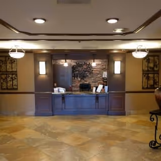 Reception desk area in an assisted living facility with a stone accent wall behind the desk, decorative wall art, and warm lighting fixtures on the ceiling and walls.