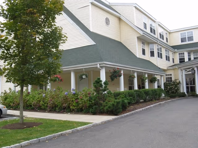 Exterior view of a multi-story senior living facility with beige siding and a green roof. The building has a covered porch with white columns and hanging flower baskets. There are shrubs, flowers, and a tree planted along the sidewalk and driveway.