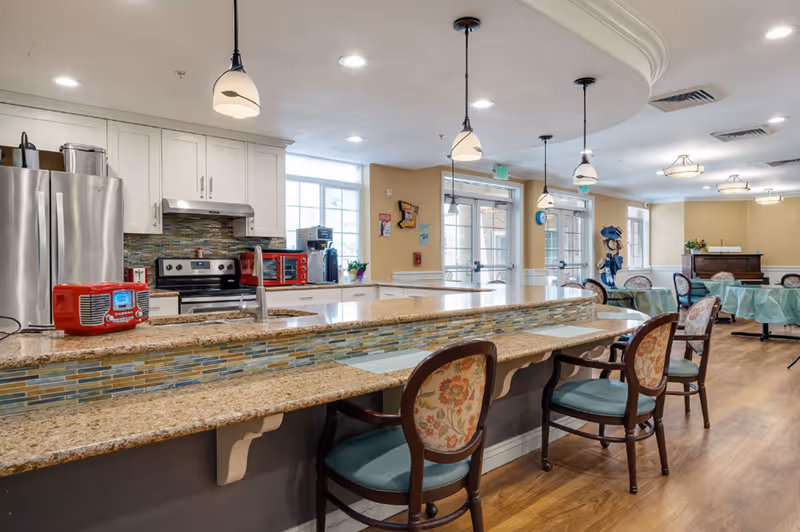 A bright and spacious kitchen and dining area in a senior living facility. The kitchen features white cabinets, stainless steel refrigerator and stove, a red toaster oven, and a red radio on the granite countertop. There are pendant lights hanging above the counter with floral upholstered chairs lined up along the counter. In the background, there are tables covered with green tablecloths and chairs, a piano, and large windows letting in natural light.
