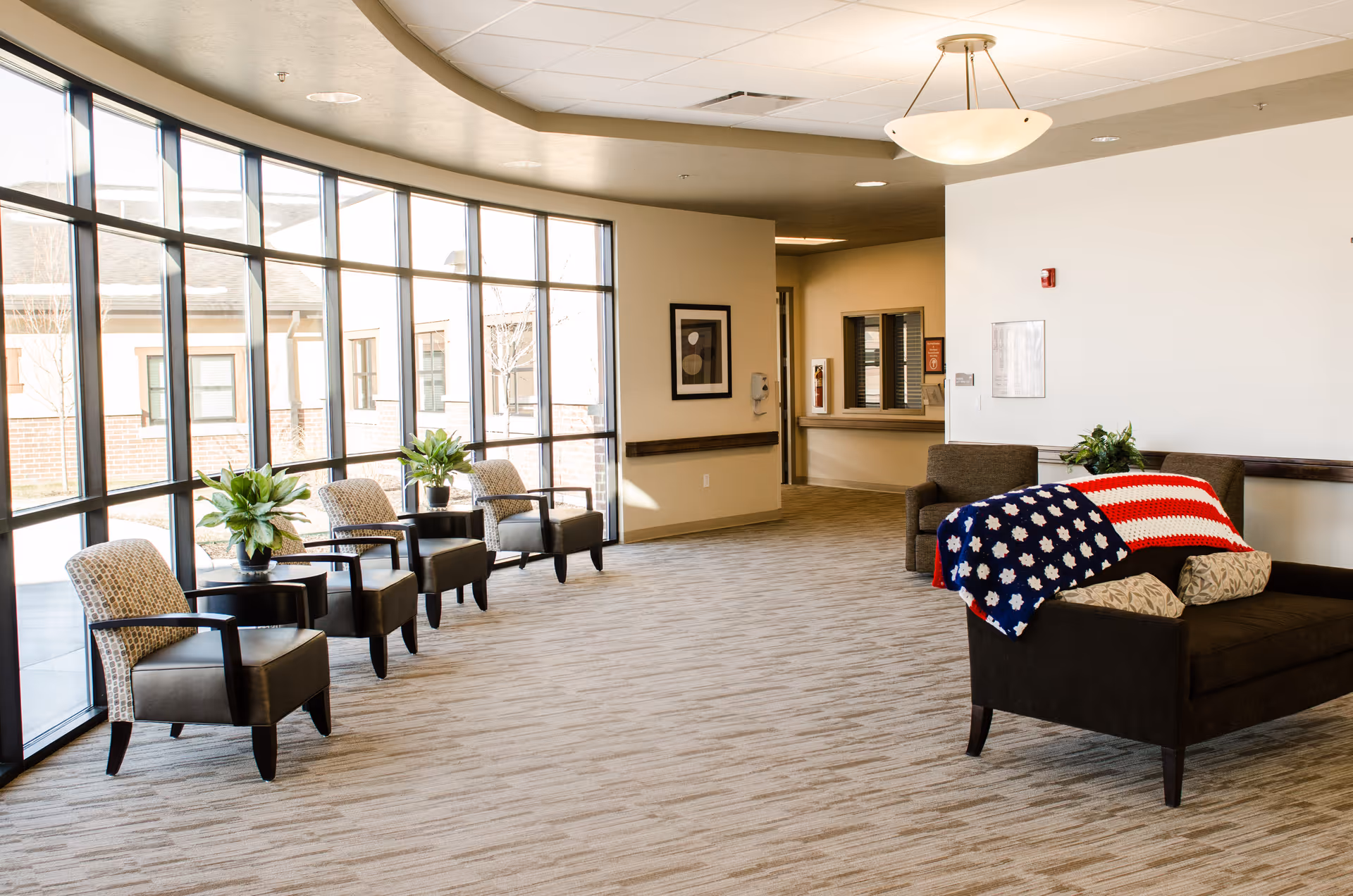 A spacious, well-lit common area with large floor-to-ceiling windows along one wall. The room features a row of armchairs with small tables and potted plants in front of the windows. On the right side, there is a dark brown sofa with an American flag-themed blanket draped over the back. The walls are light-colored, and the ceiling has a modern light fixture. There is a framed picture on the wall and a reception window in the background.