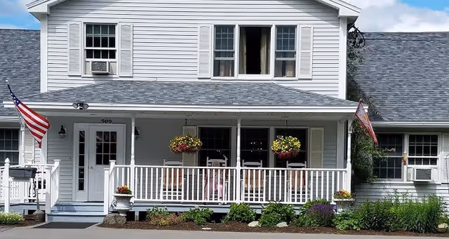 Front exterior view of a two-story white house with a porch featuring hanging flower baskets, rocking chairs, and an American flag on the left side. The house has multiple windows and a gray shingled roof.