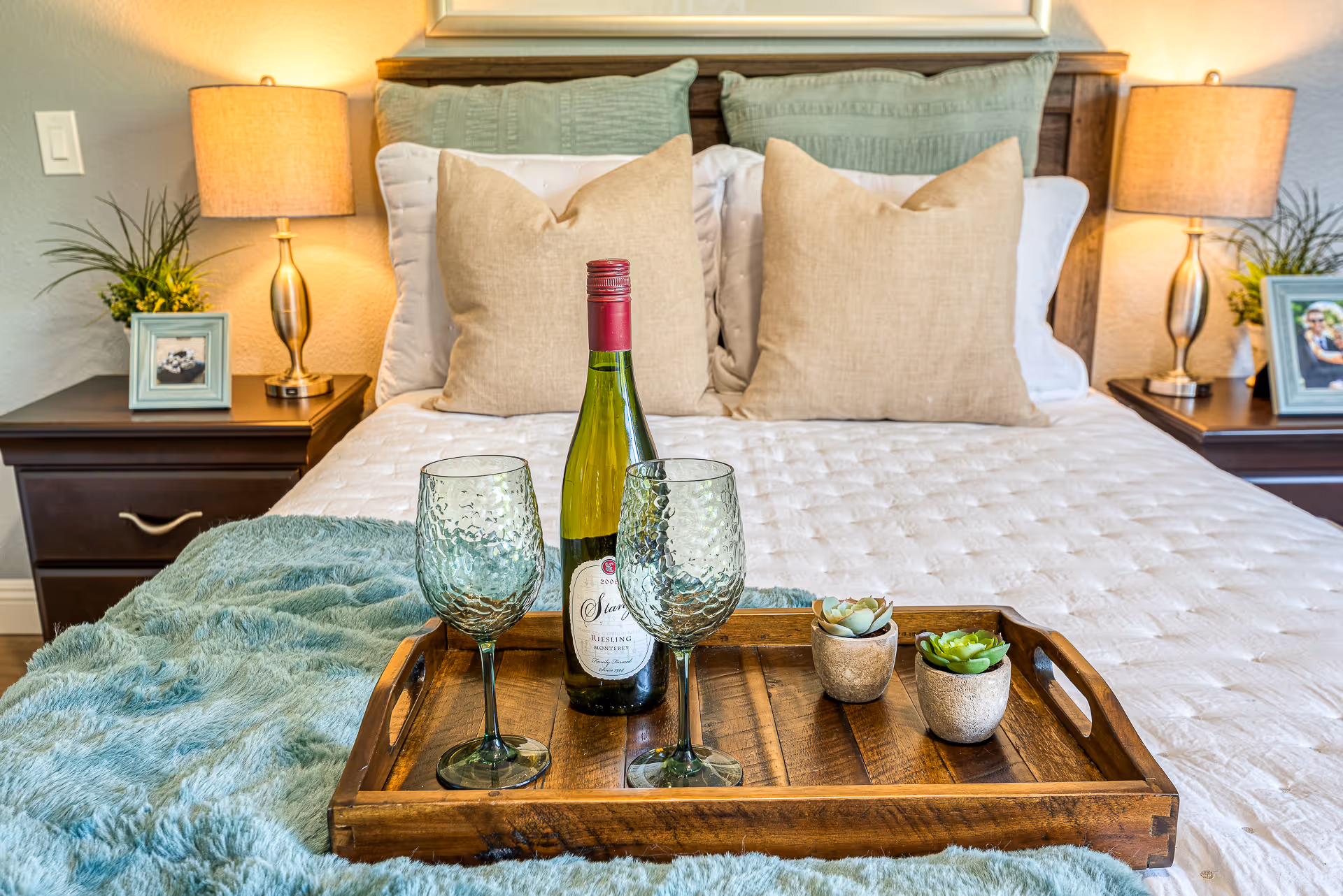 A neatly made bed with beige and green pillows, two bedside tables with lamps, framed photos, and plants. On the bed is a wooden tray holding a bottle of white wine, two textured wine glasses, and two small potted succulents.