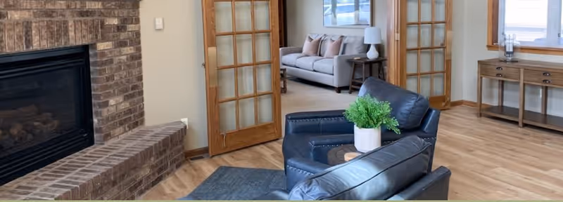 Cozy living room with leather armchairs, a brick fireplace, and glass-paned French doors opening to a sofa in an adjacent room.