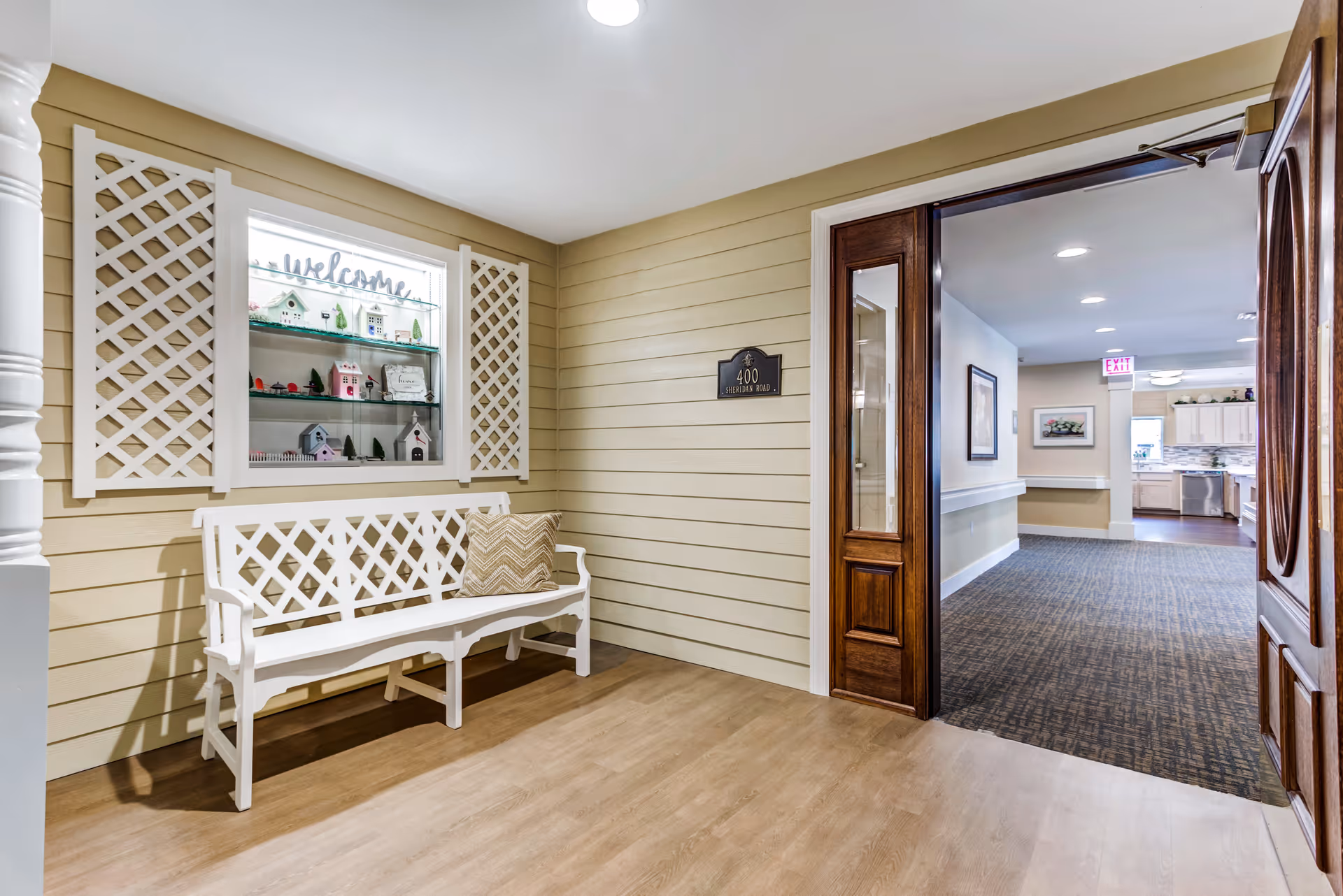 A cozy interior corner with a white wooden bench featuring a decorative pillow, set against a beige wall with white lattice panels and a glass display case containing miniature houses and a 'welcome' sign. To the right, a wooden door opens into a hallway leading to a kitchen area with white cabinets and stainless steel appliances.